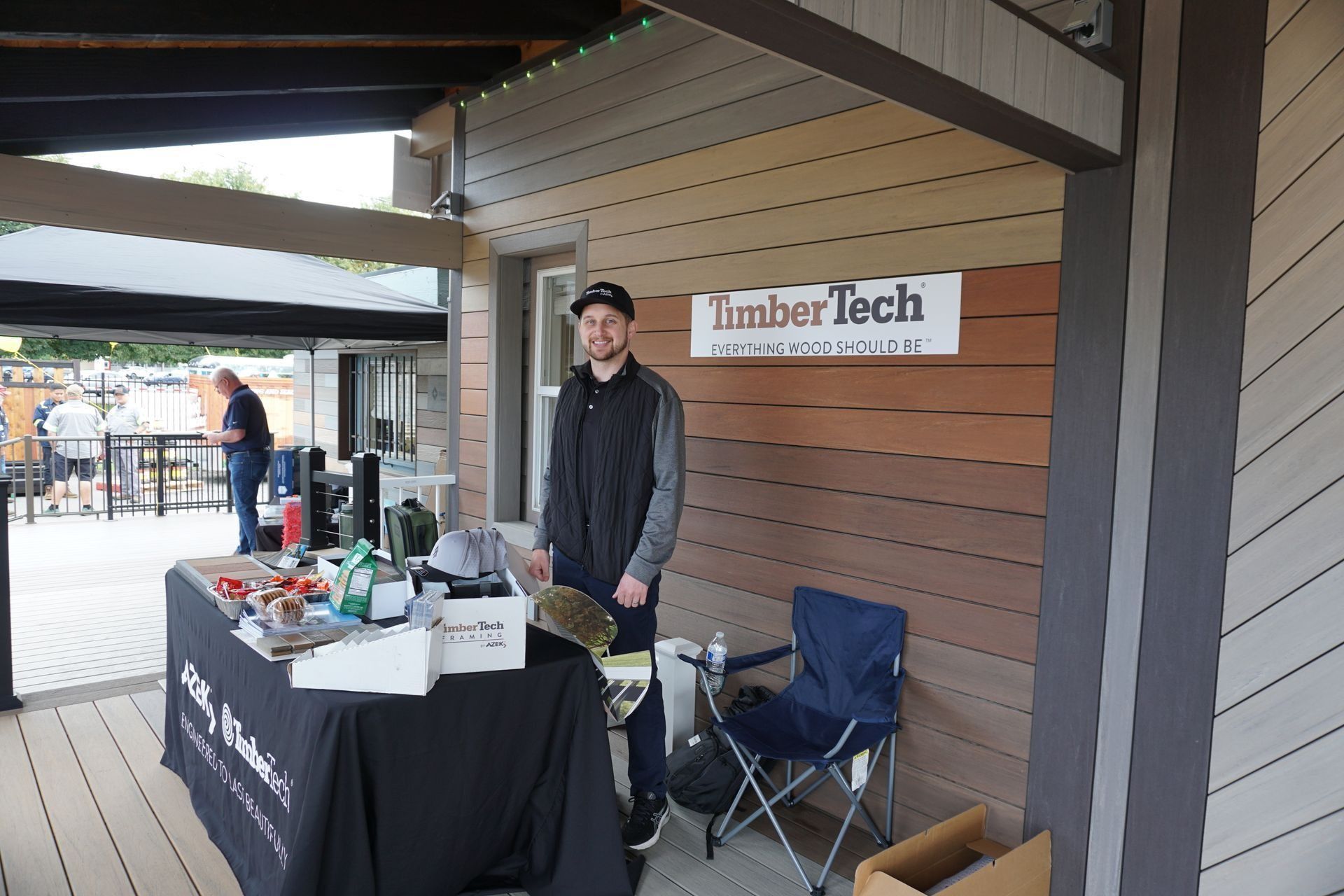 Man stands at a TimberTech booth outdoors, holding a skateboard. Table with product samples. Brown and beige building.