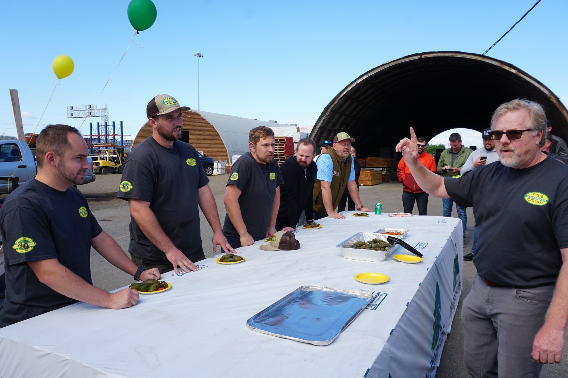 People at table lined up, eating competition. Man gestures, others watch, balloons in background.