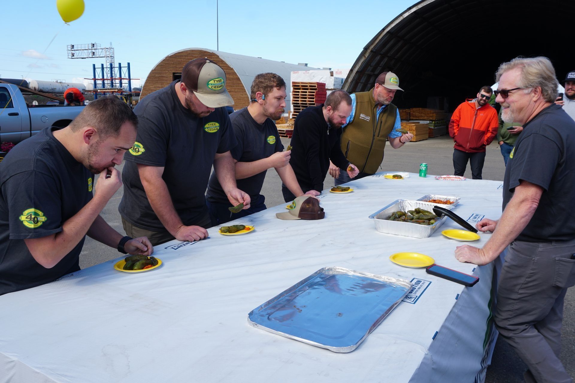 Men in matching shirts compete in an outdoor eating contest at a table laden with food.