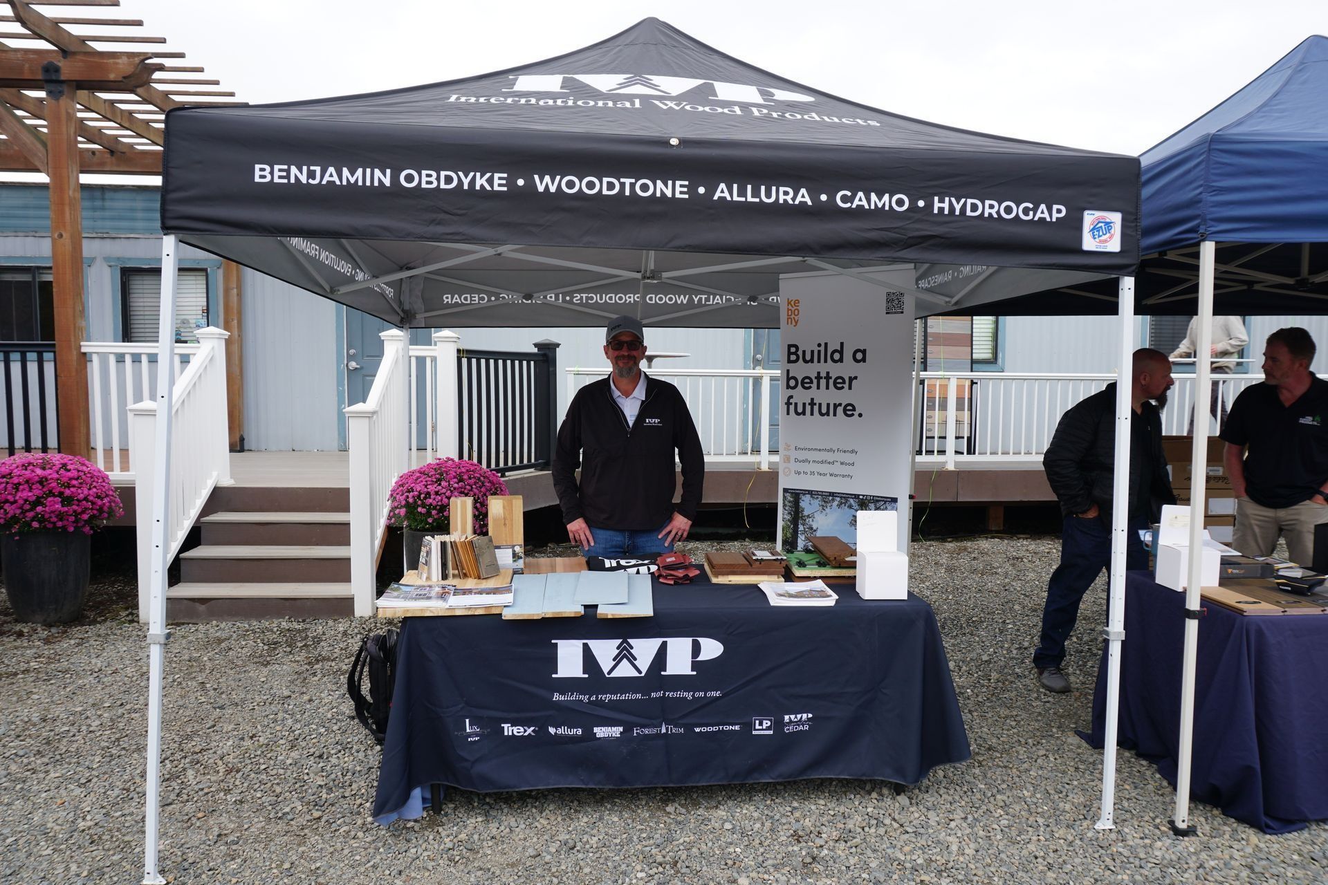IWP booth with man, display table, product samples, and canopy. Outdoor setting with blue and white building in background.