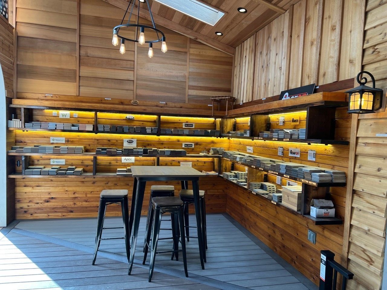 Wooden-walled retail space with shelves displaying products, a small table with stools, and a decorative light fixture.