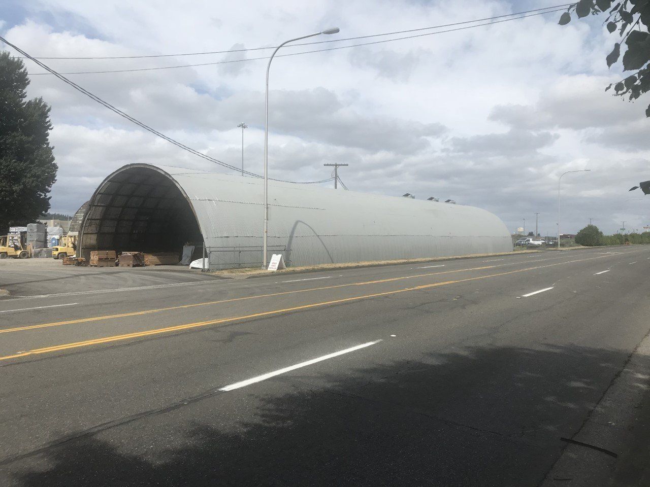 Large gray arched building next to a paved road under a cloudy sky.