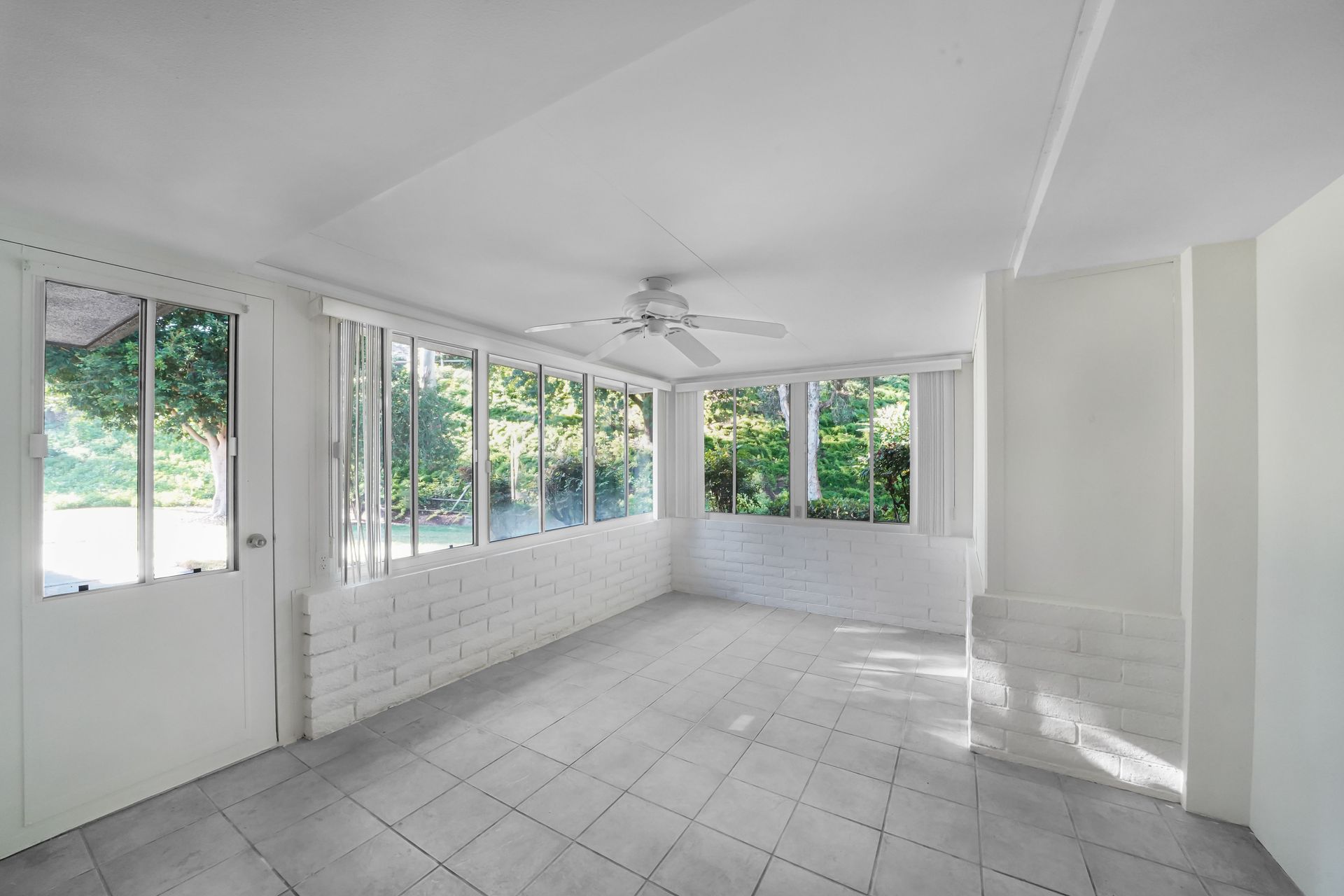 Bright empty sunroom with white tiled floor, large windows, and a ceiling fan.