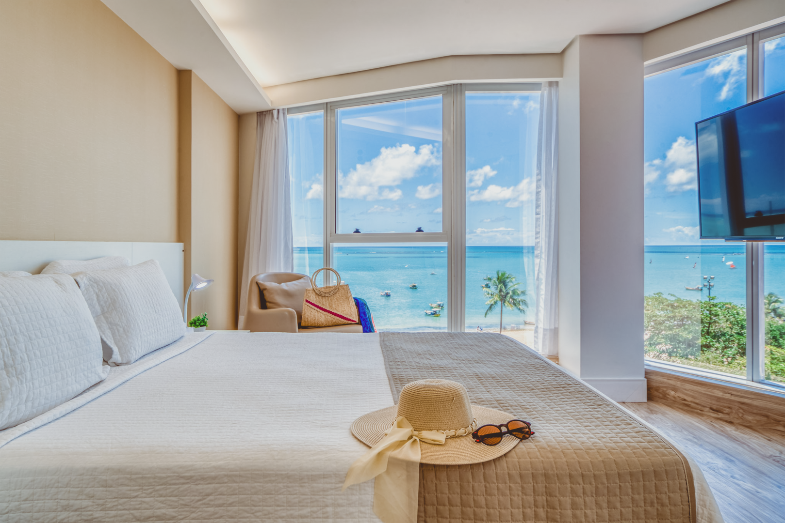 Quarto com cama, chapéu e vista para o mar através de grandes janelas.