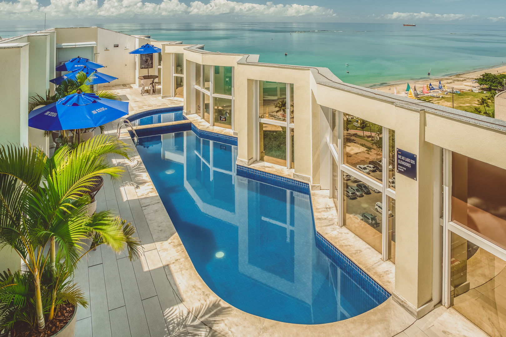 Piscina com vista para o mar, prédios brancos com guarda-sóis azuis, palmeiras.