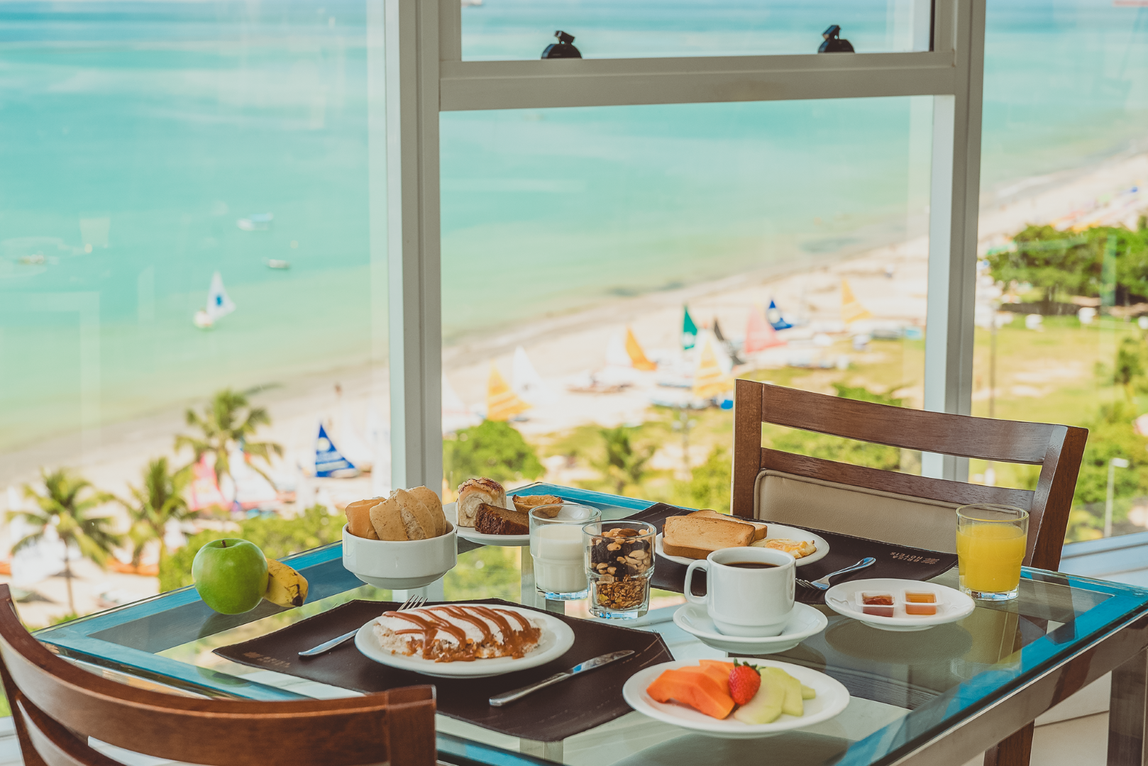 Mesa de café da manhã perto de uma janela com vista para a praia, com frutas, doces e café.