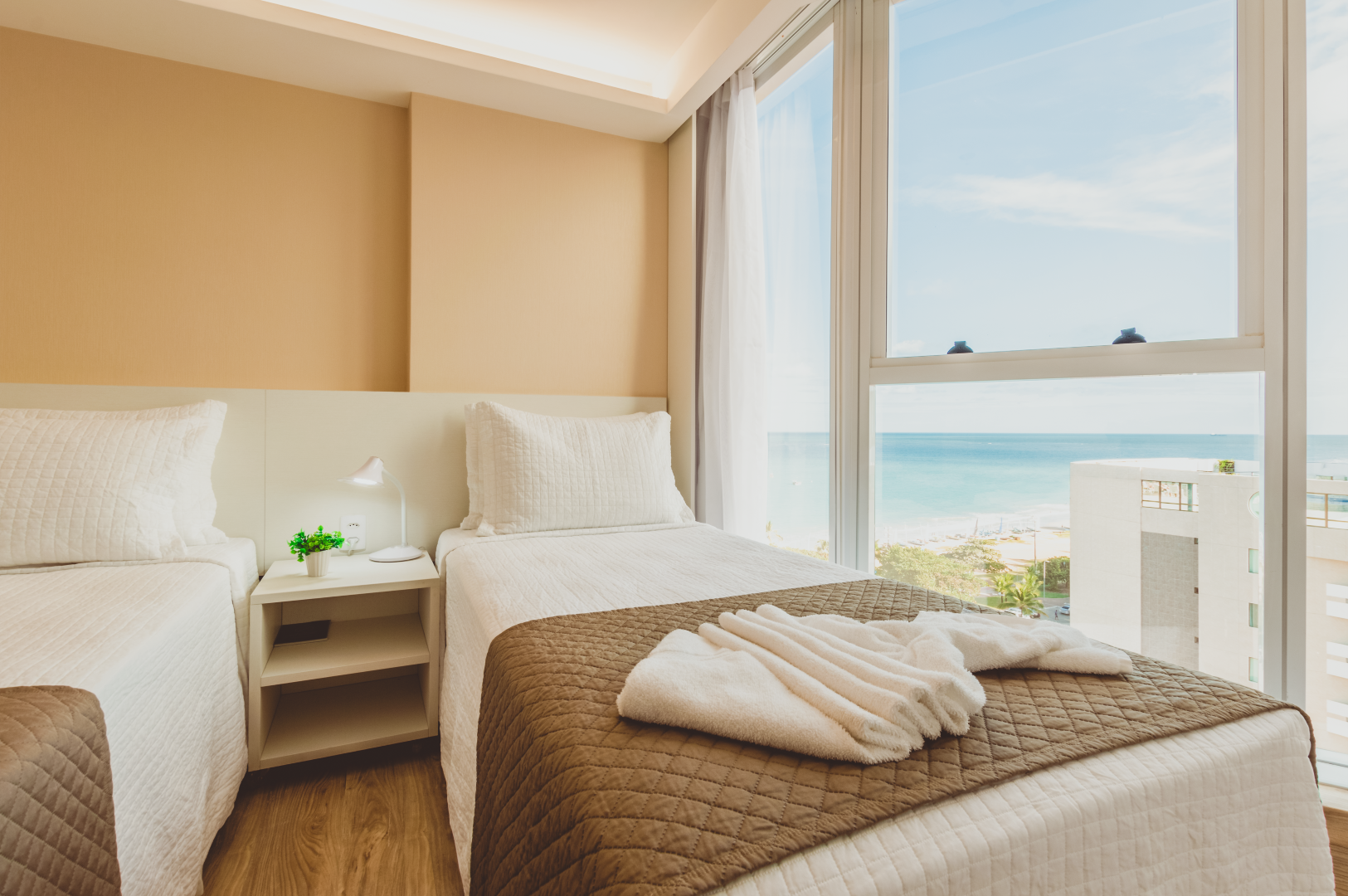 Duas camas de solteiro em um quarto bege com vista para o mar. Lençóis brancos, edredom marrom e mesa de cabeceira pequena.