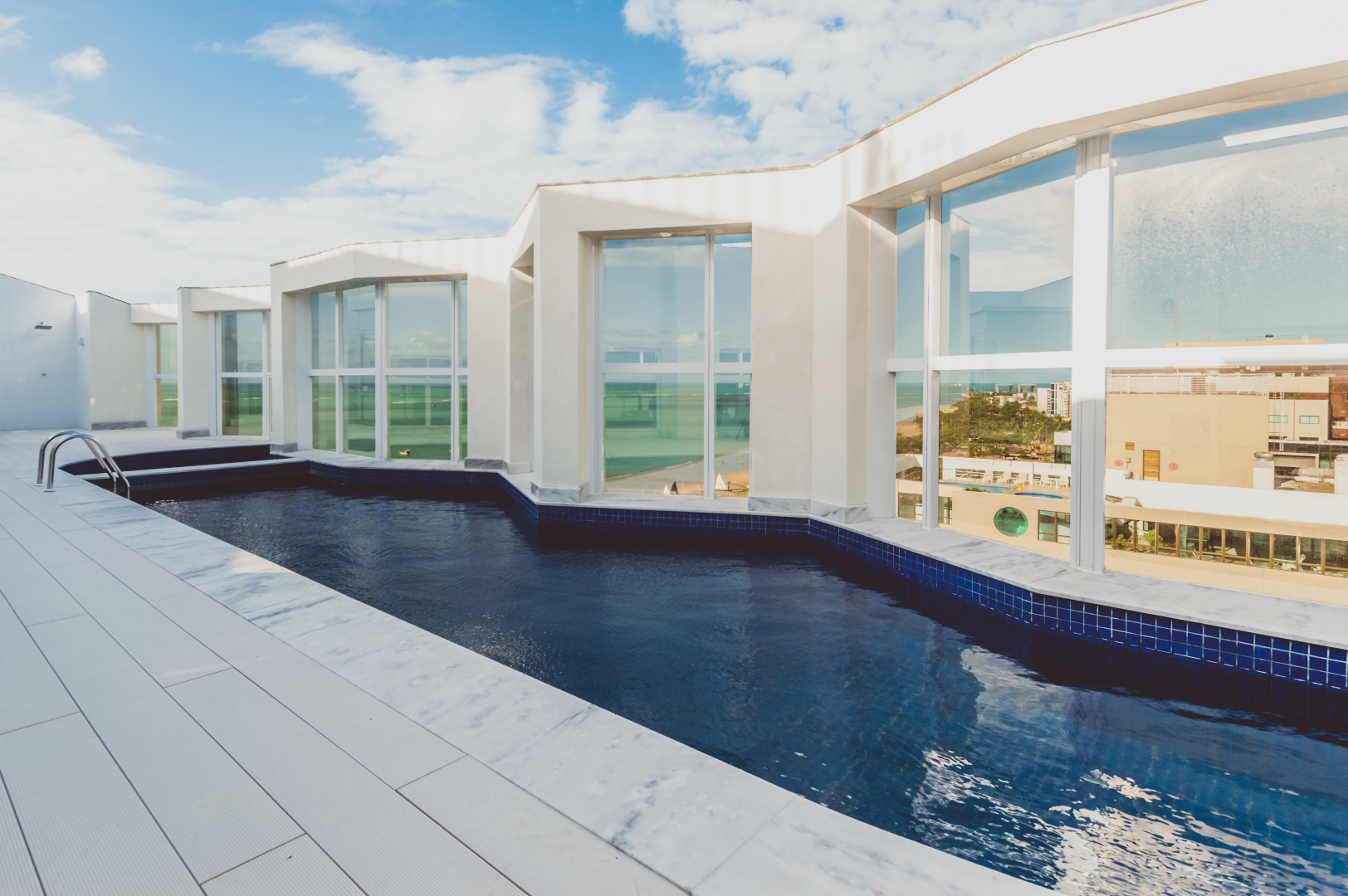 Piscina na cobertura com azulejos brancos e água azul, cercada por um prédio branco com grandes janelas, dia ensolarado.