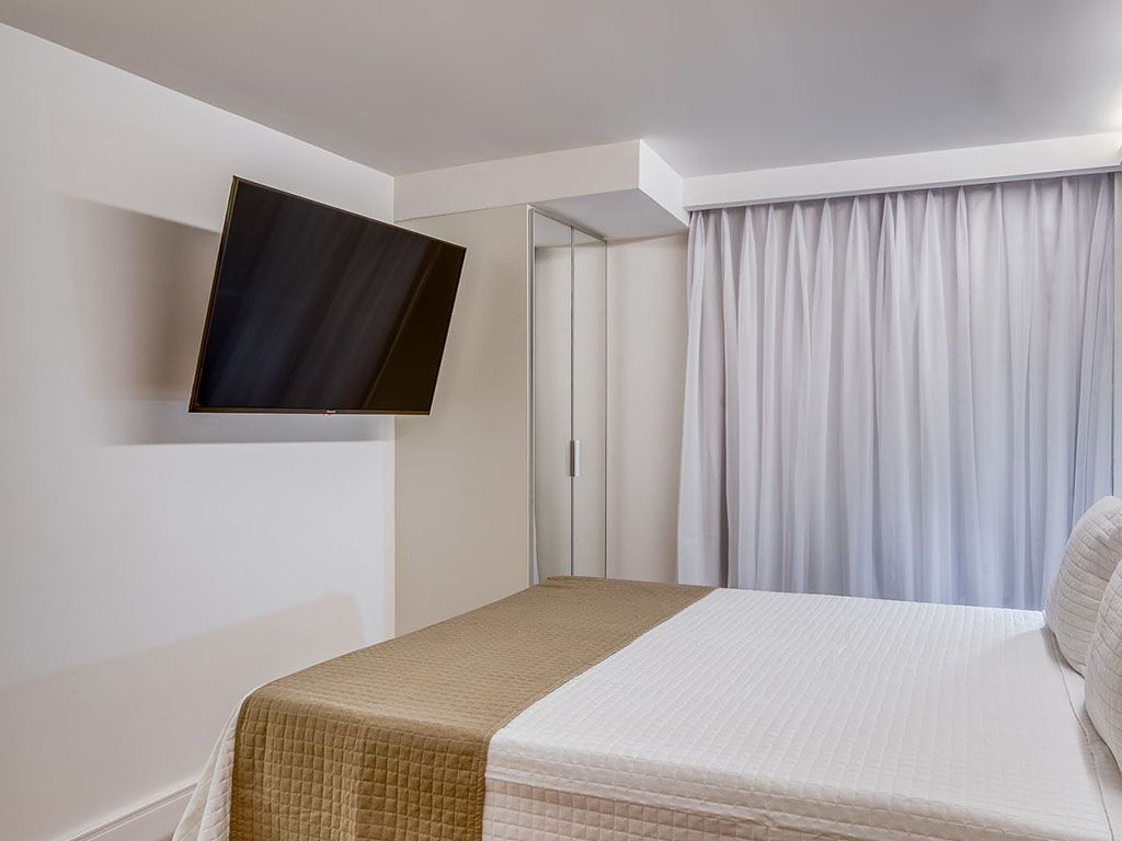 Quarto com TV na parede, cama com roupa de cama branca e bege e cortinas brancas.