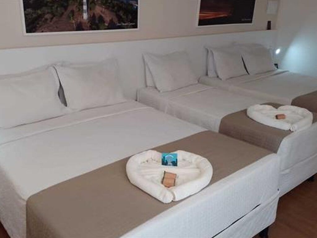 Três camas de hotel com lençóis brancos, passadeiras cinza-claro e toalhas em formato de coração.