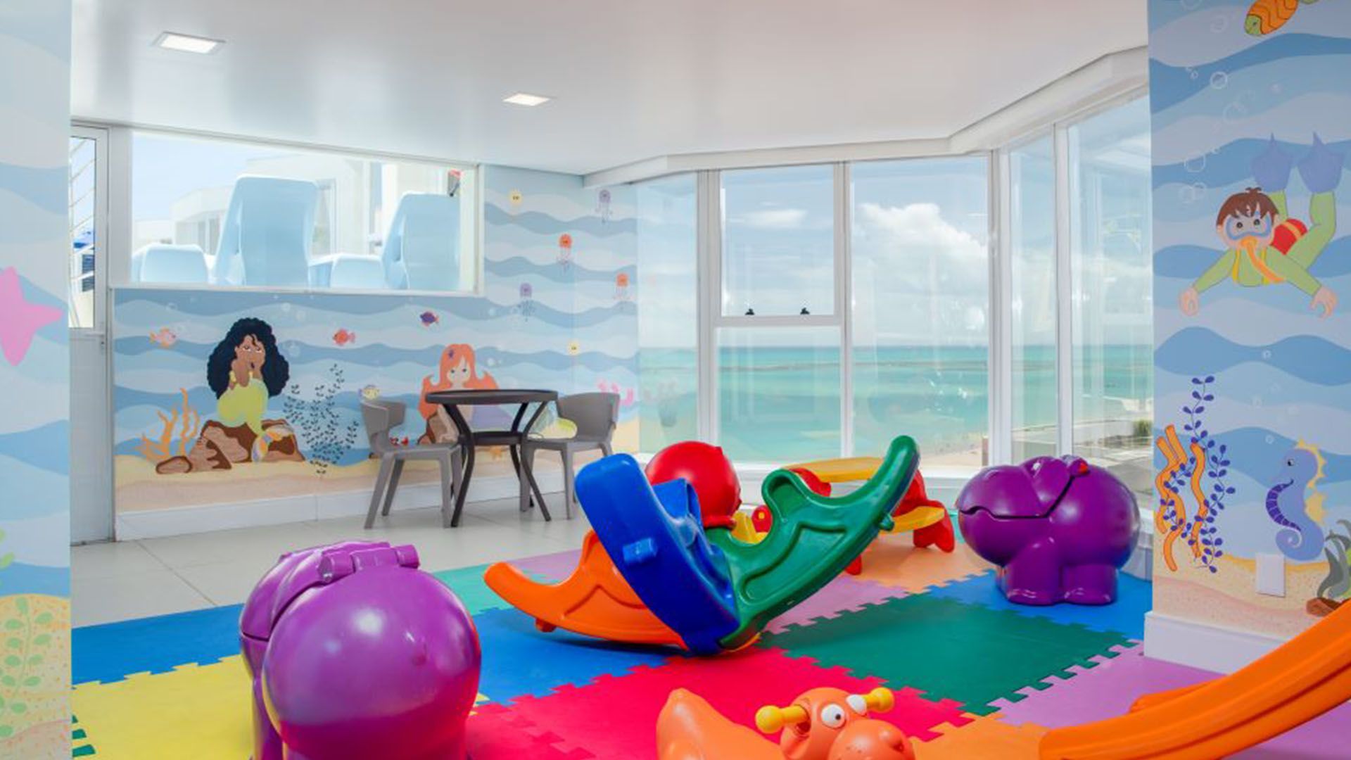 Sala de jogos com vista para o mar, brinquedos coloridos, murais e uma pequena mesa.