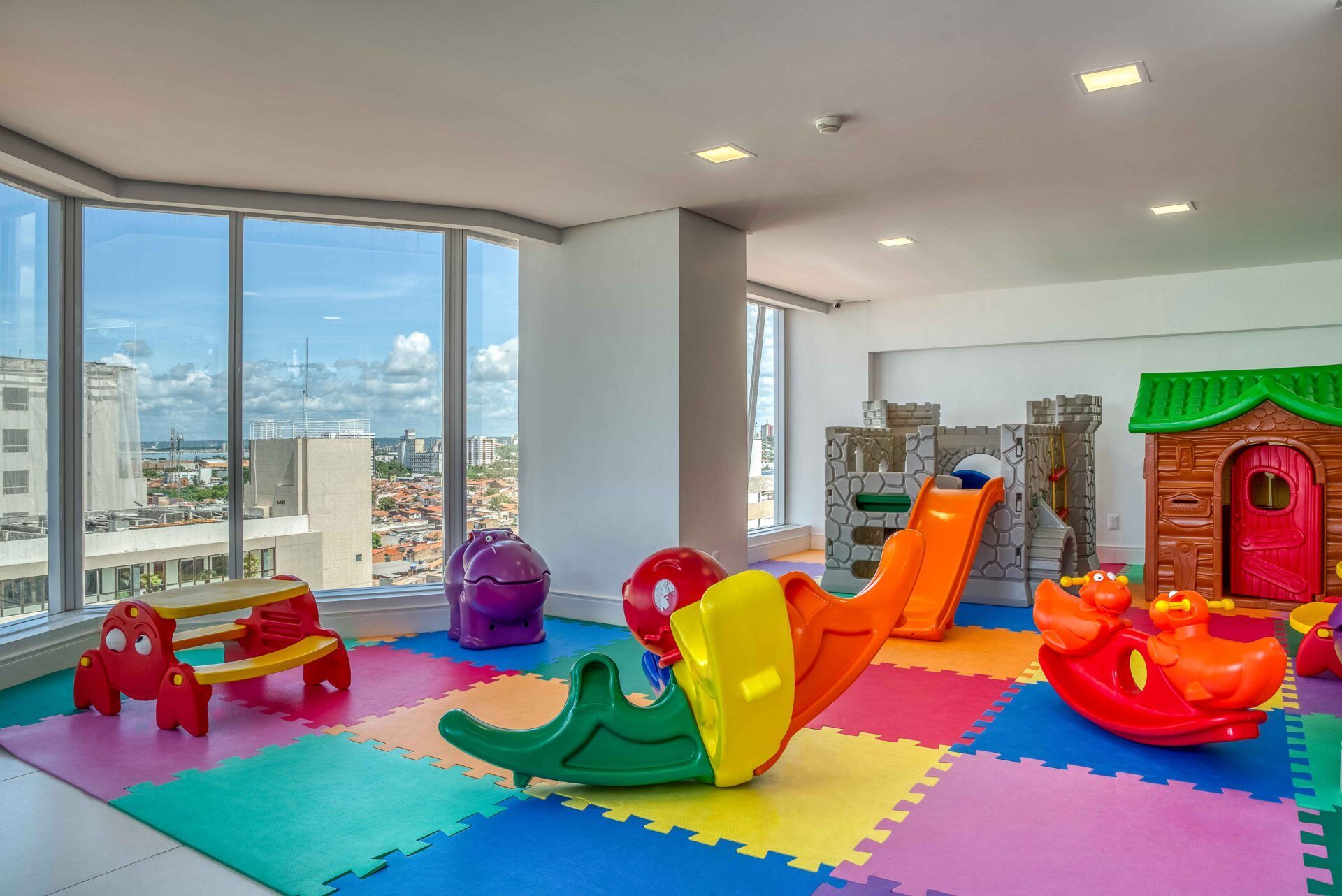 Sala de jogos com piso acolchoado colorido, brinquedos, escorregador em formato de castelo e grandes janelas com vista para a cidade.