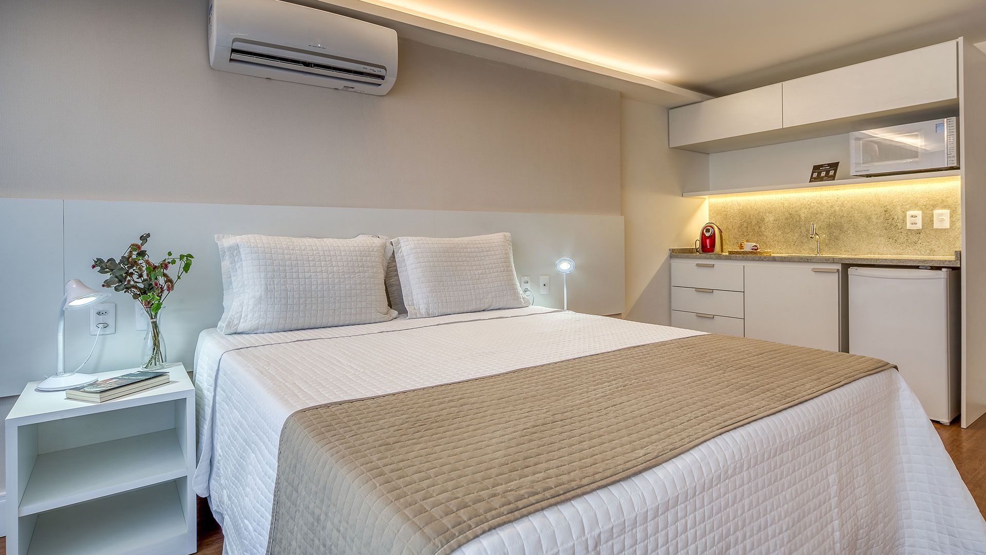 Quarto branco com cama, mesa de cabeceira, cozinha compacta e ar condicionado.