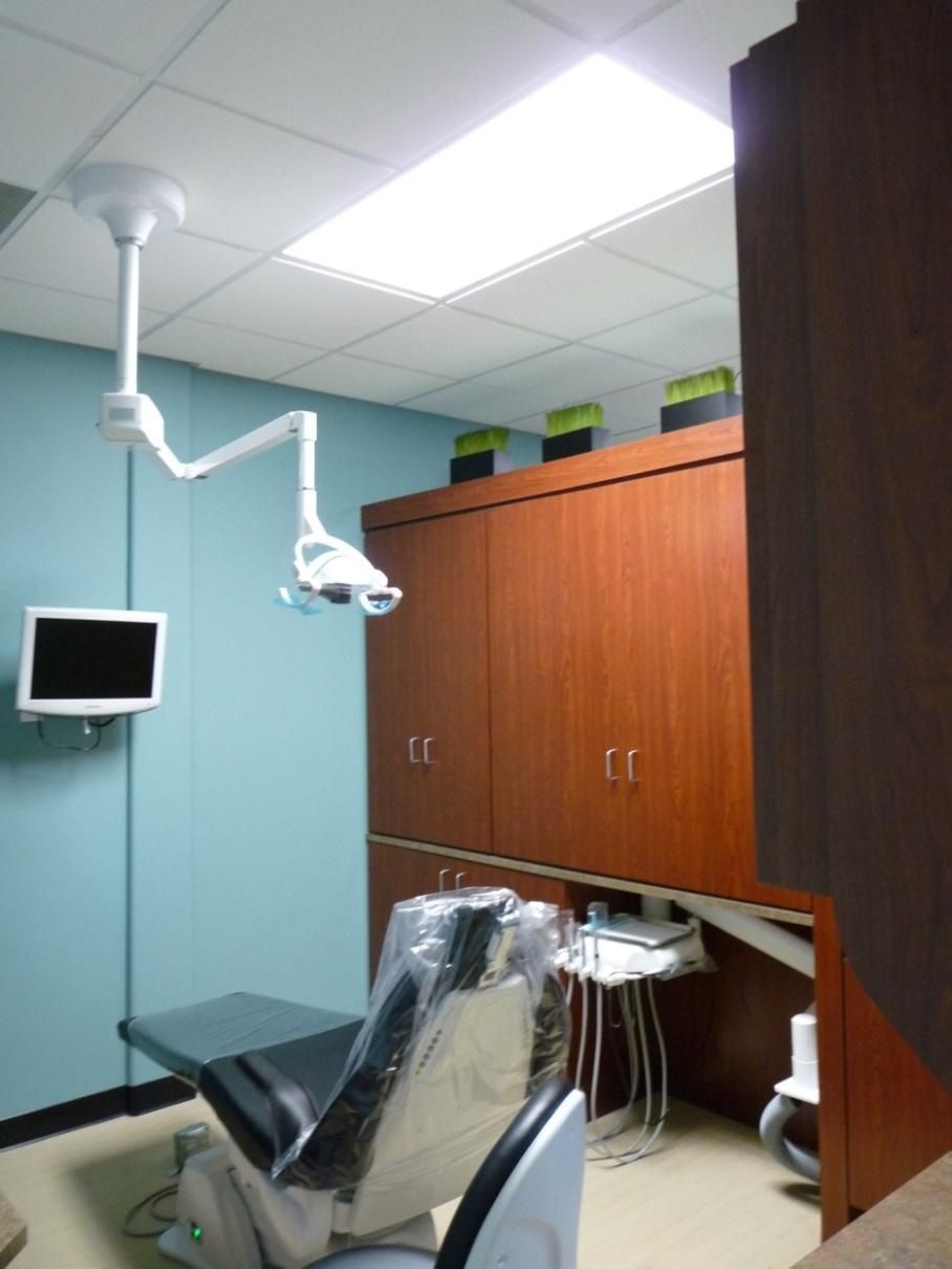 Dental exam room: chair, light, cabinets, monitor, and tools. Blue walls, wood cabinets, and a bright ceiling light.