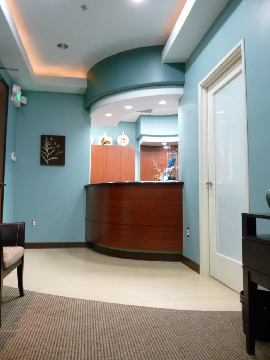 Reception area in a medical office. Teal walls, wood desk, beige floor, and a closed white door.