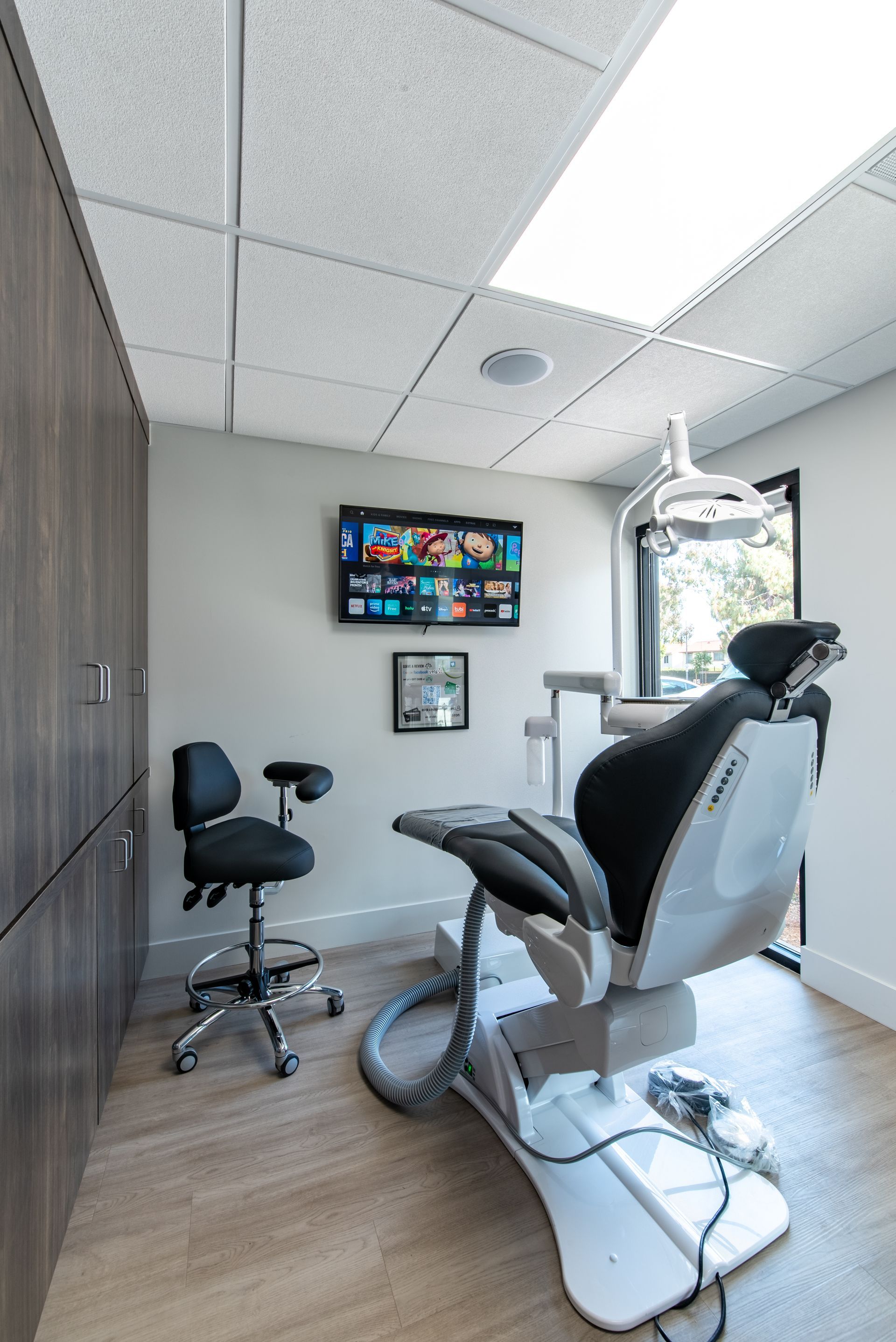 Dental office: Patient chair, black stool, cabinetry, TV, window. Beige walls and flooring.