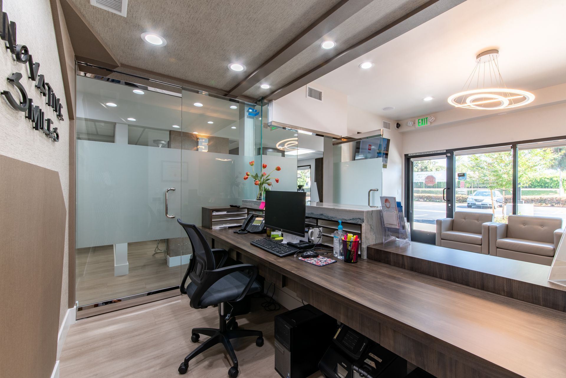Reception area with a desk, computer, and chairs, near a glass-walled office.