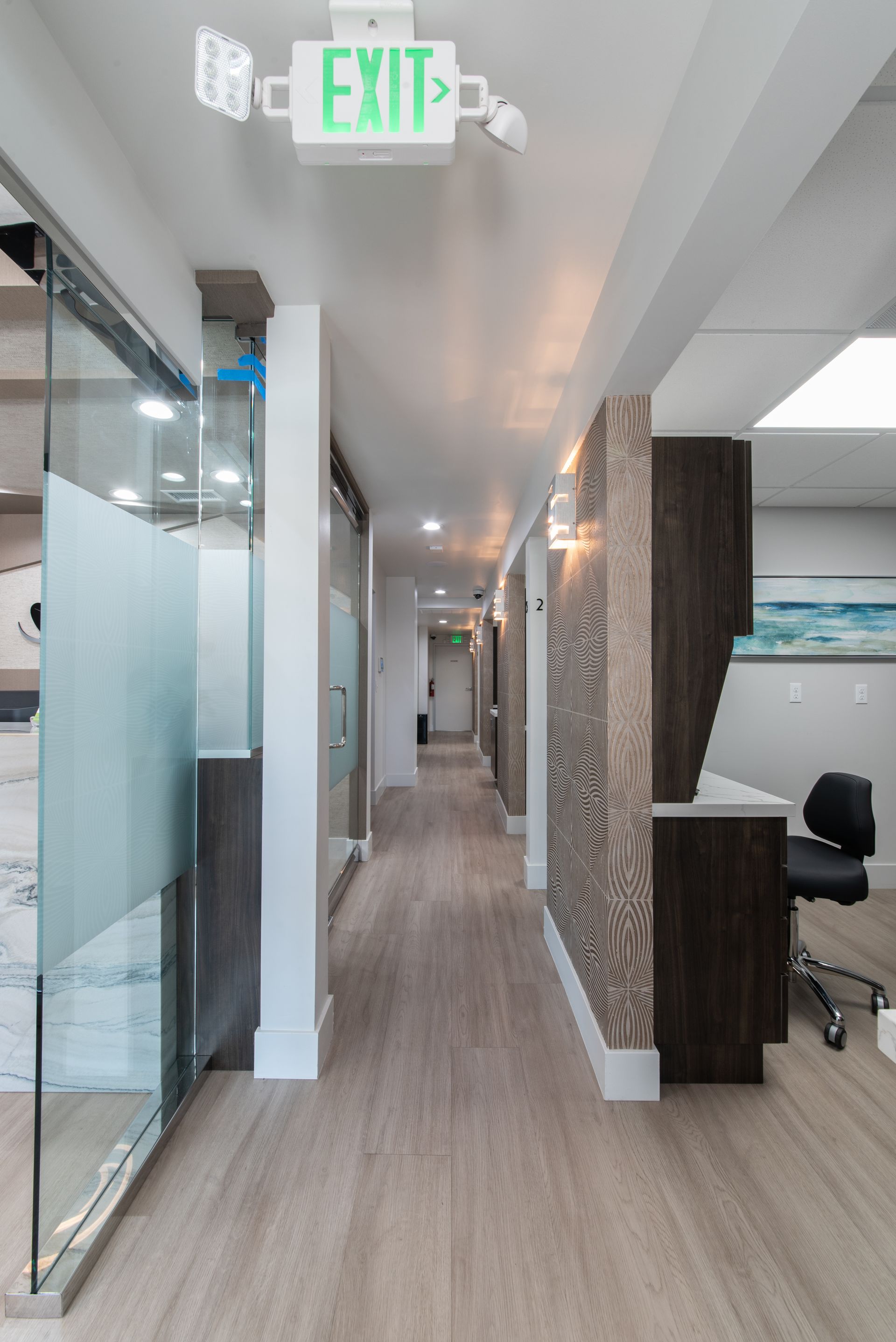 Hallway in a medical office with wood-look flooring, glass doors, and an exit sign.