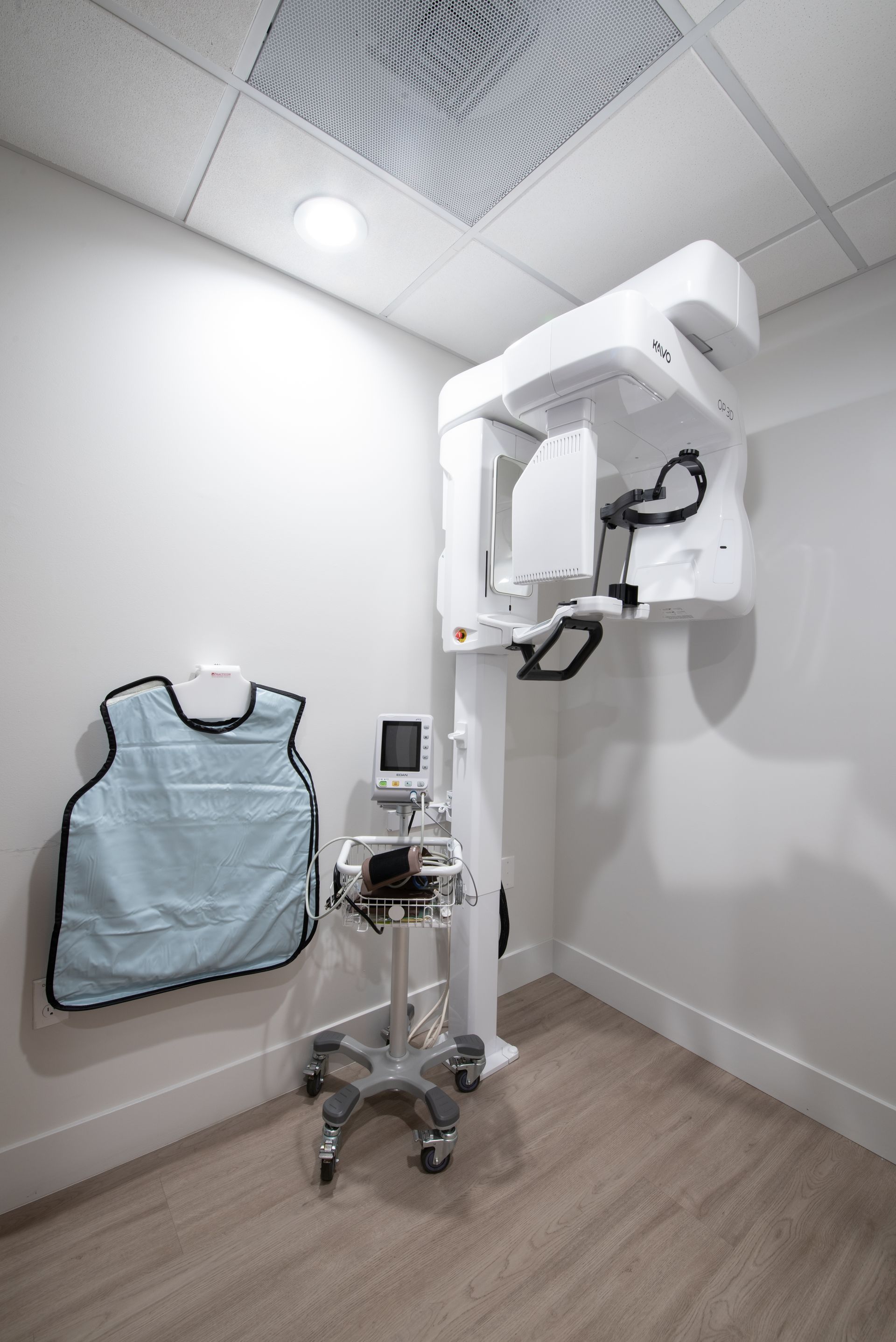 Dental X-ray machine in a corner of a white room, with a lead apron hanging on the wall.