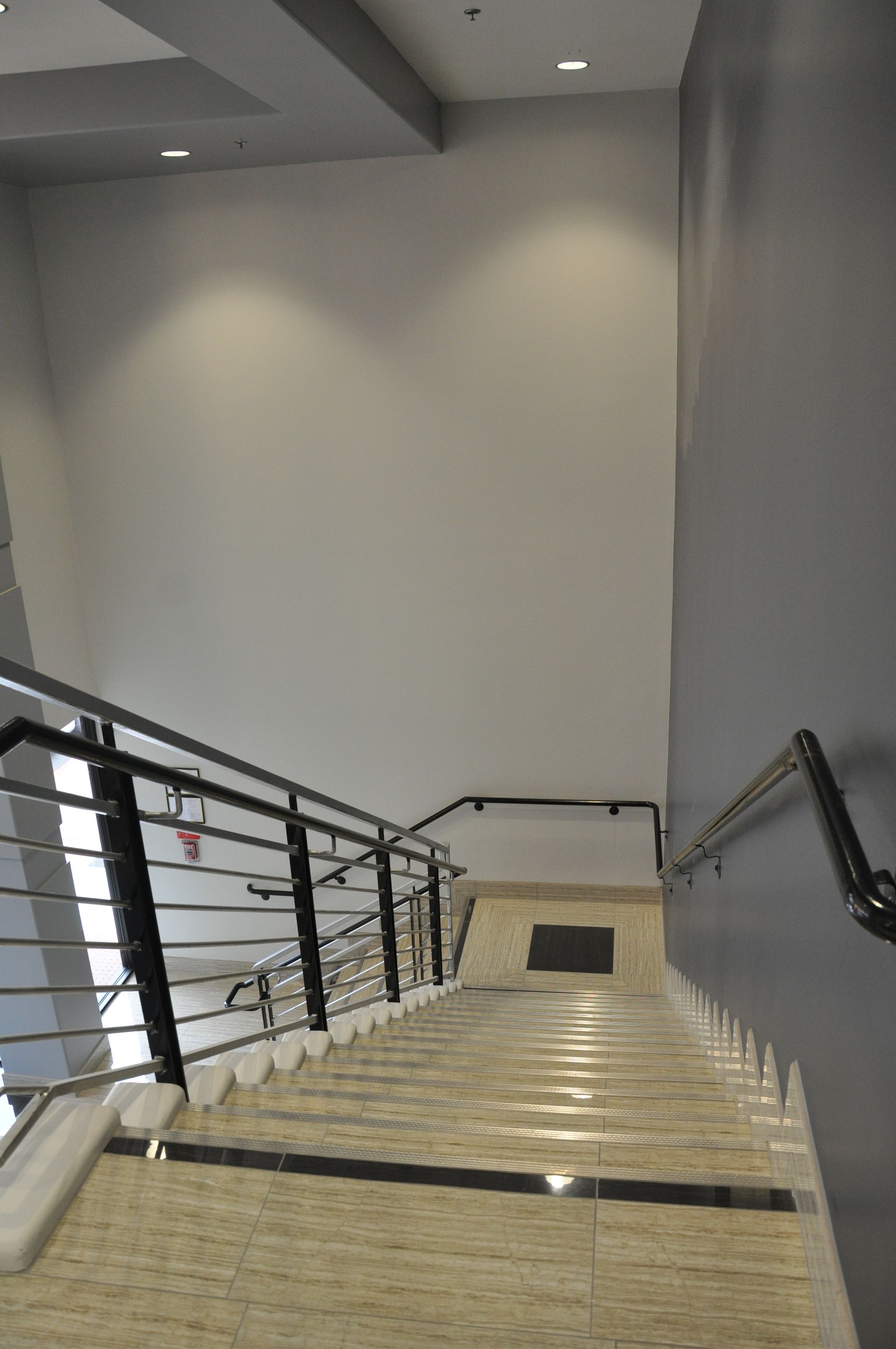 Stairwell leading down. Beige tile steps, black and silver railings, gray walls.