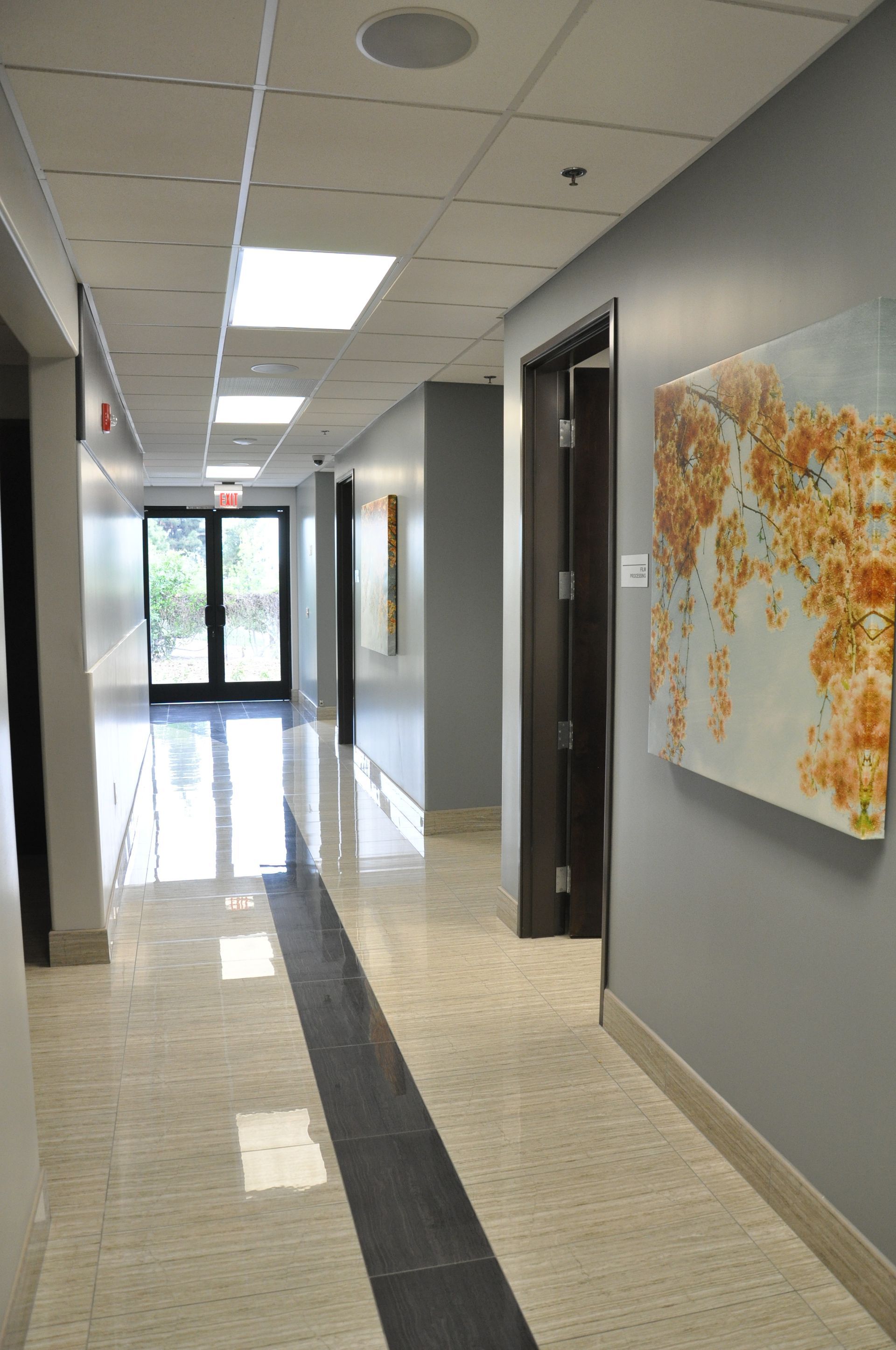 Hallway with gray walls, tile floor, and artwork, leading to glass doors and natural light.