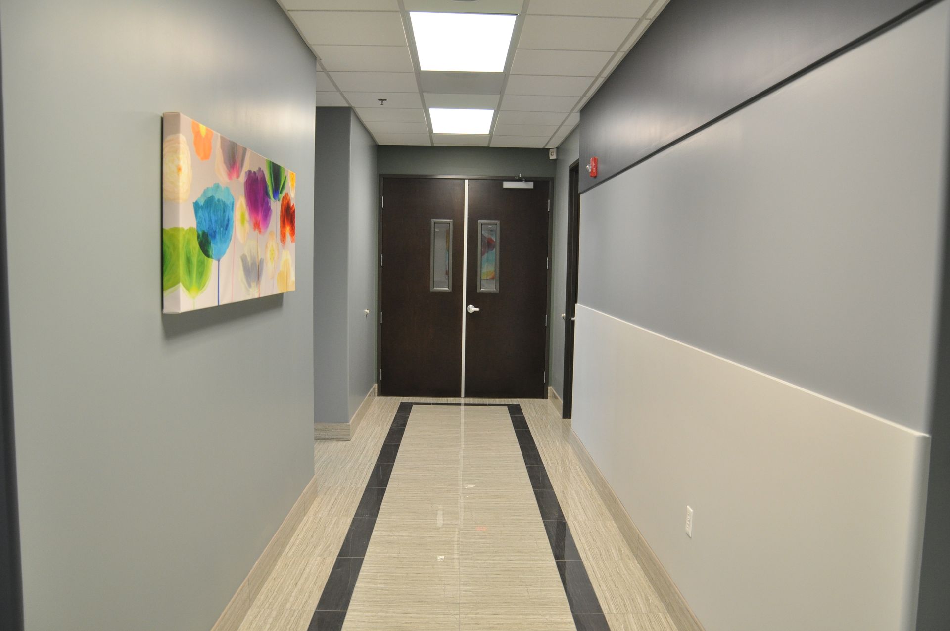 Narrow hallway with gray and white walls, art, and closed double doors at the end.
