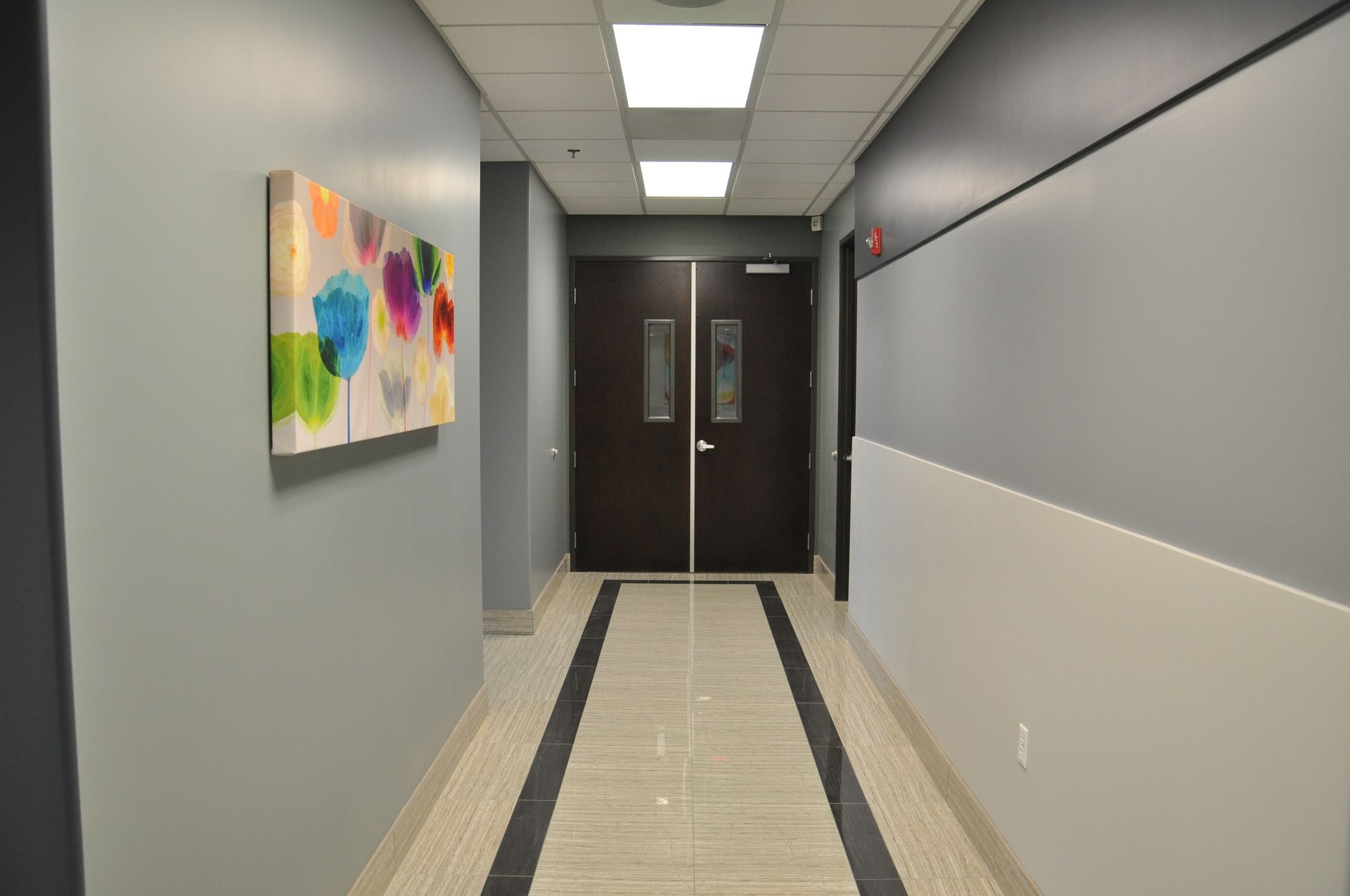 Hallway with gray and white walls, a colorful painting, and dark double doors at the end.