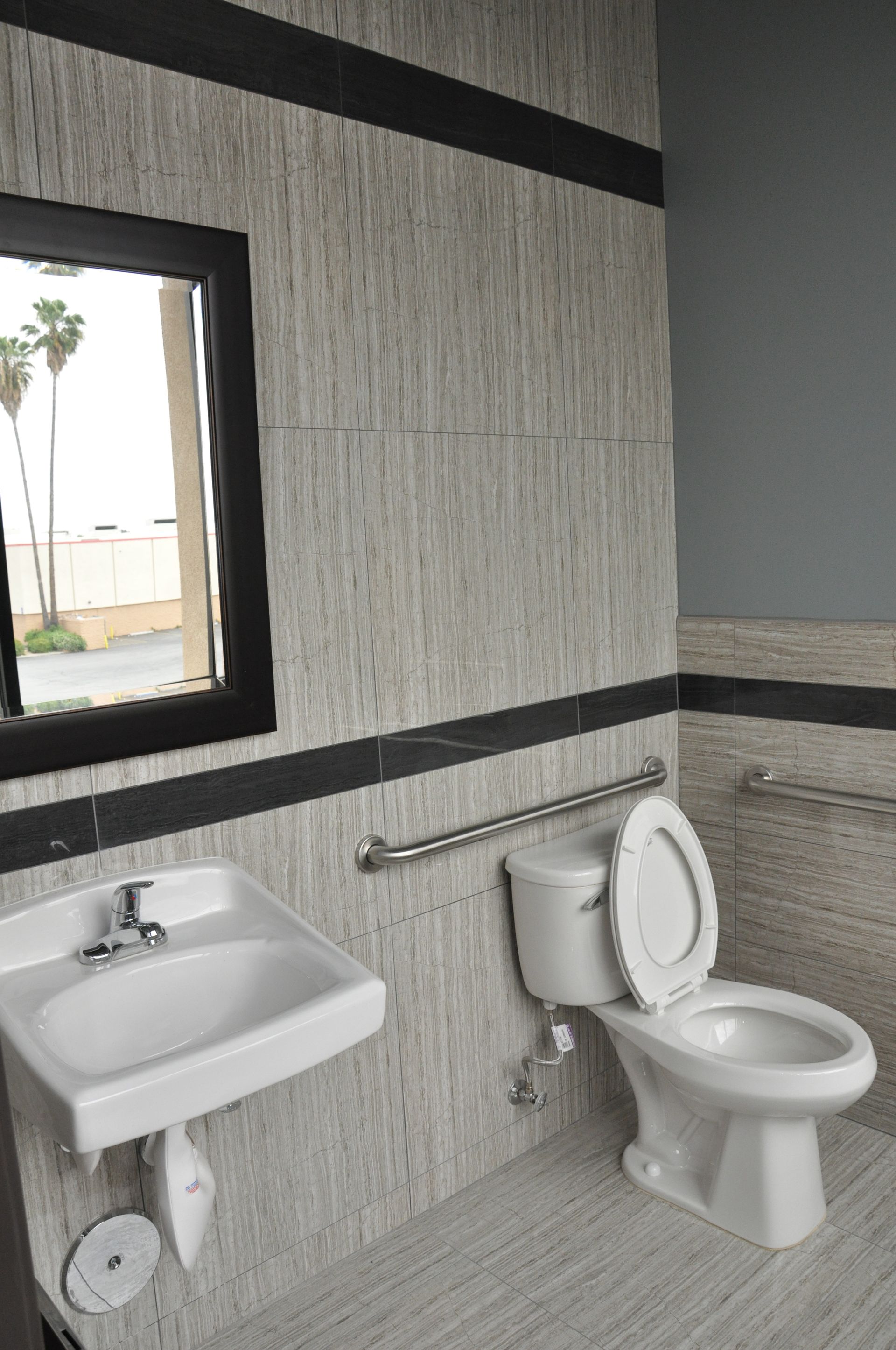 Accessible bathroom with sink, toilet, and handrails. Gray and white tiled walls, black trim.