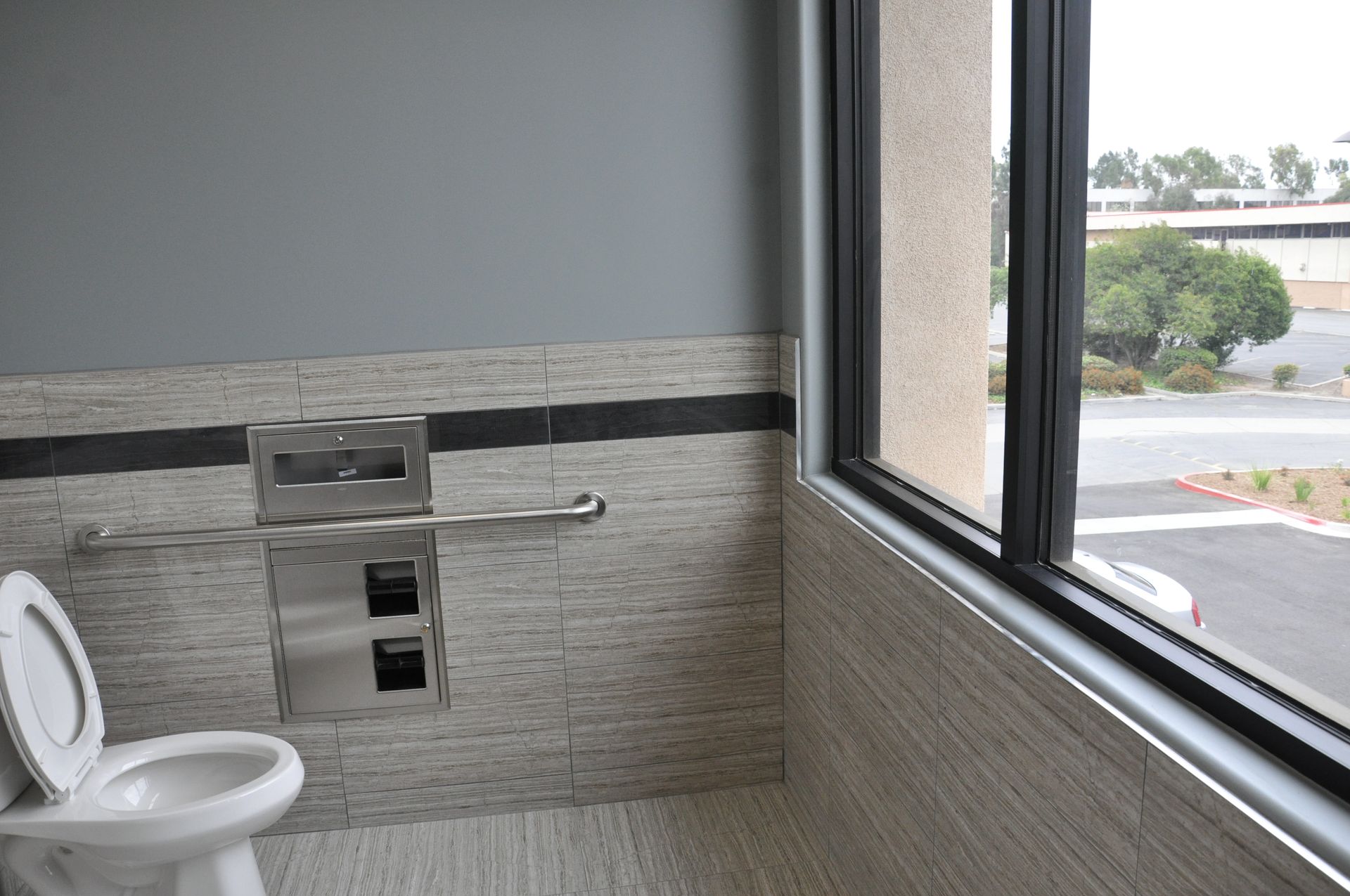 Accessible bathroom stall with toilet, handrail, paper towel dispenser, and window overlooking outdoors.