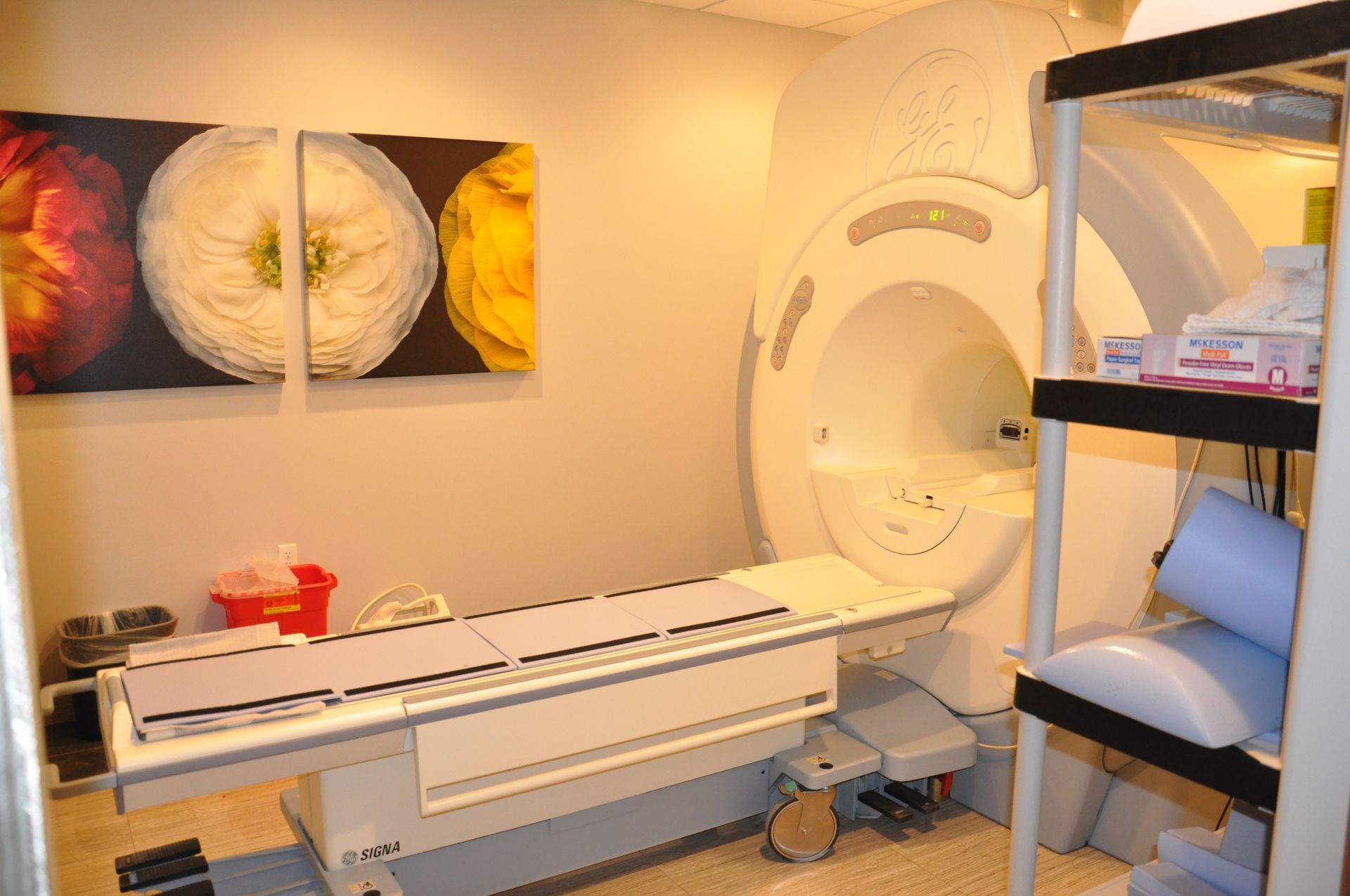 MRI machine in a medical setting with patient table and shelving with supplies. Artwork on wall.