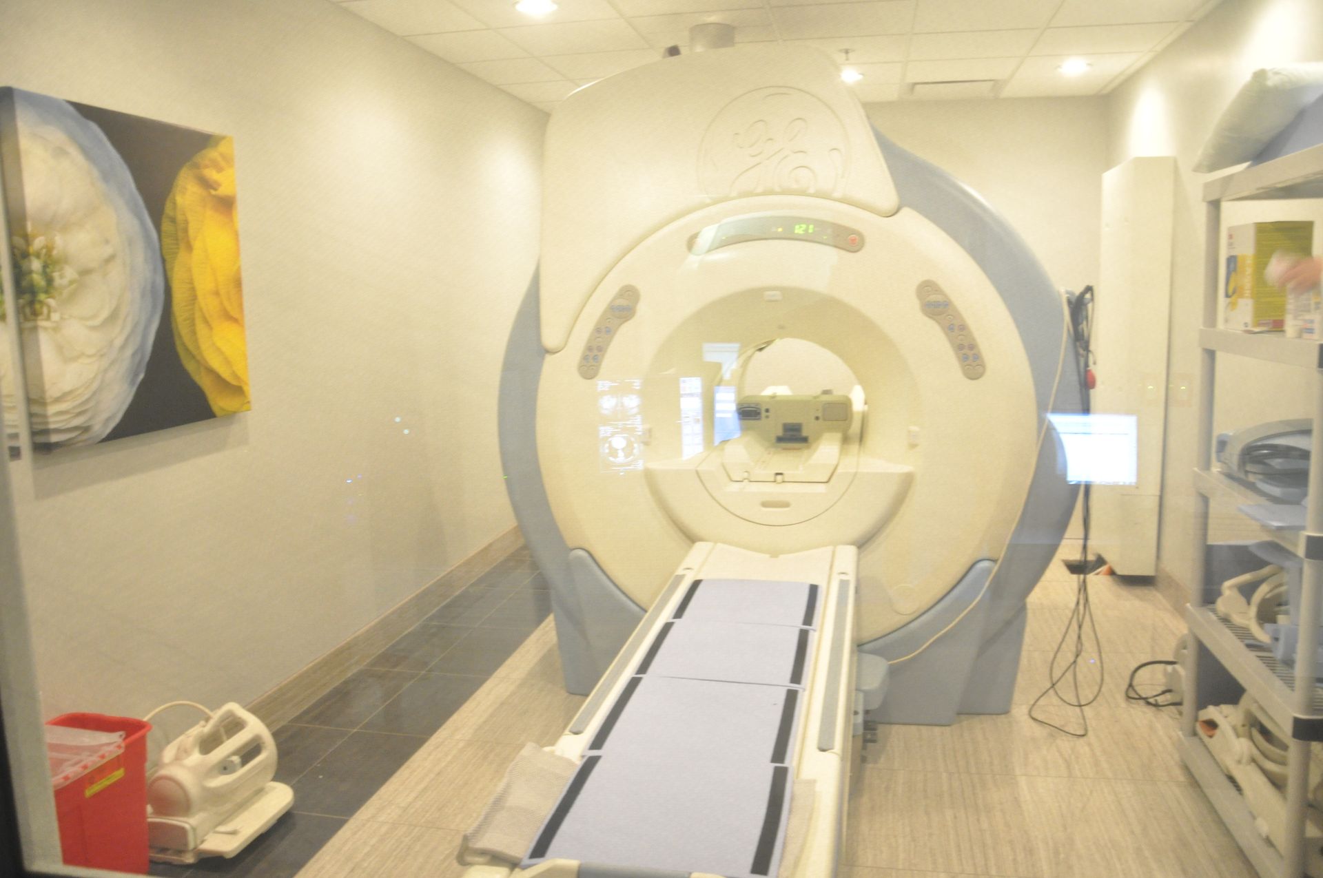 MRI machine in a clinical setting, with a patient bed, artwork on the wall and a shelving unit.