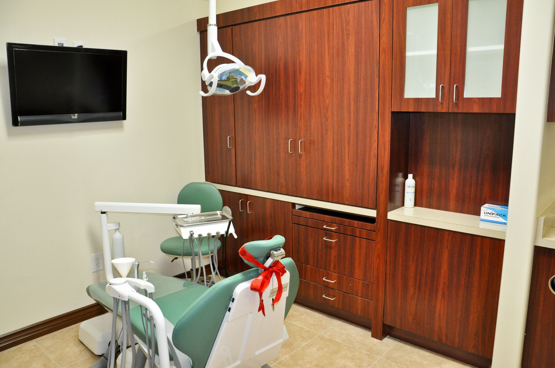 Dental office with green chair, dental light, brown cabinets, TV, and a decorative red bow.