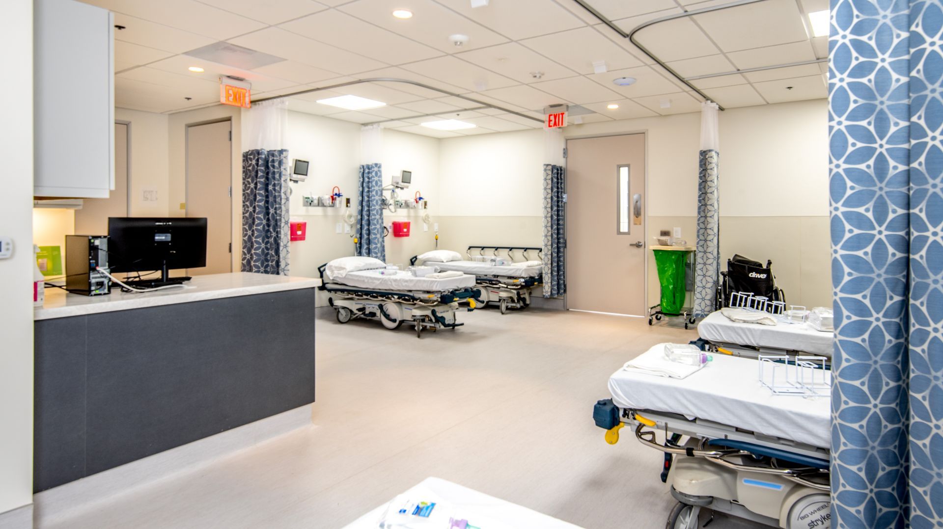 An emergency room with beds, equipment, and curtains. White walls, blue patterned curtains, and beige floors.