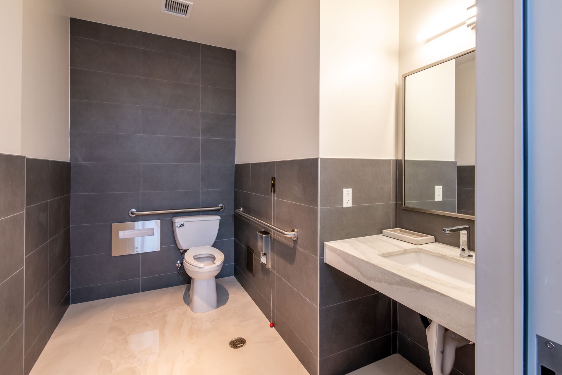 Accessible restroom with gray tile walls, white toilet, and sink. Features grab bars.