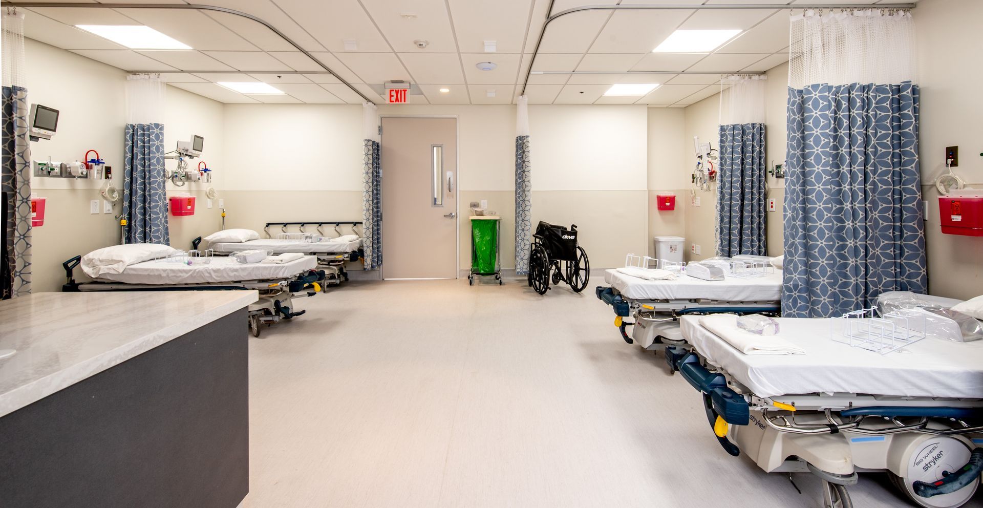 Hospital room with multiple beds, medical equipment, and blue patterned privacy curtains.