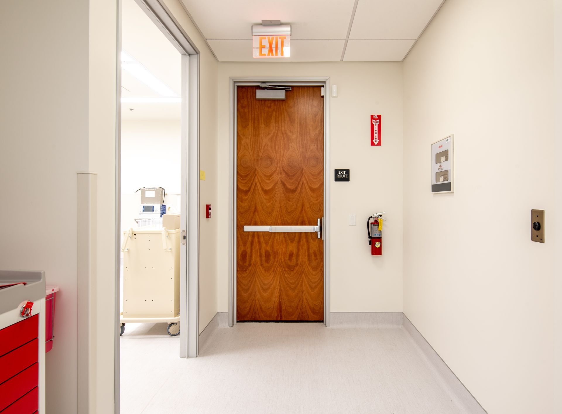 Hallway with wooden door, exit sign, fire extinguisher, and a medical cart visible.