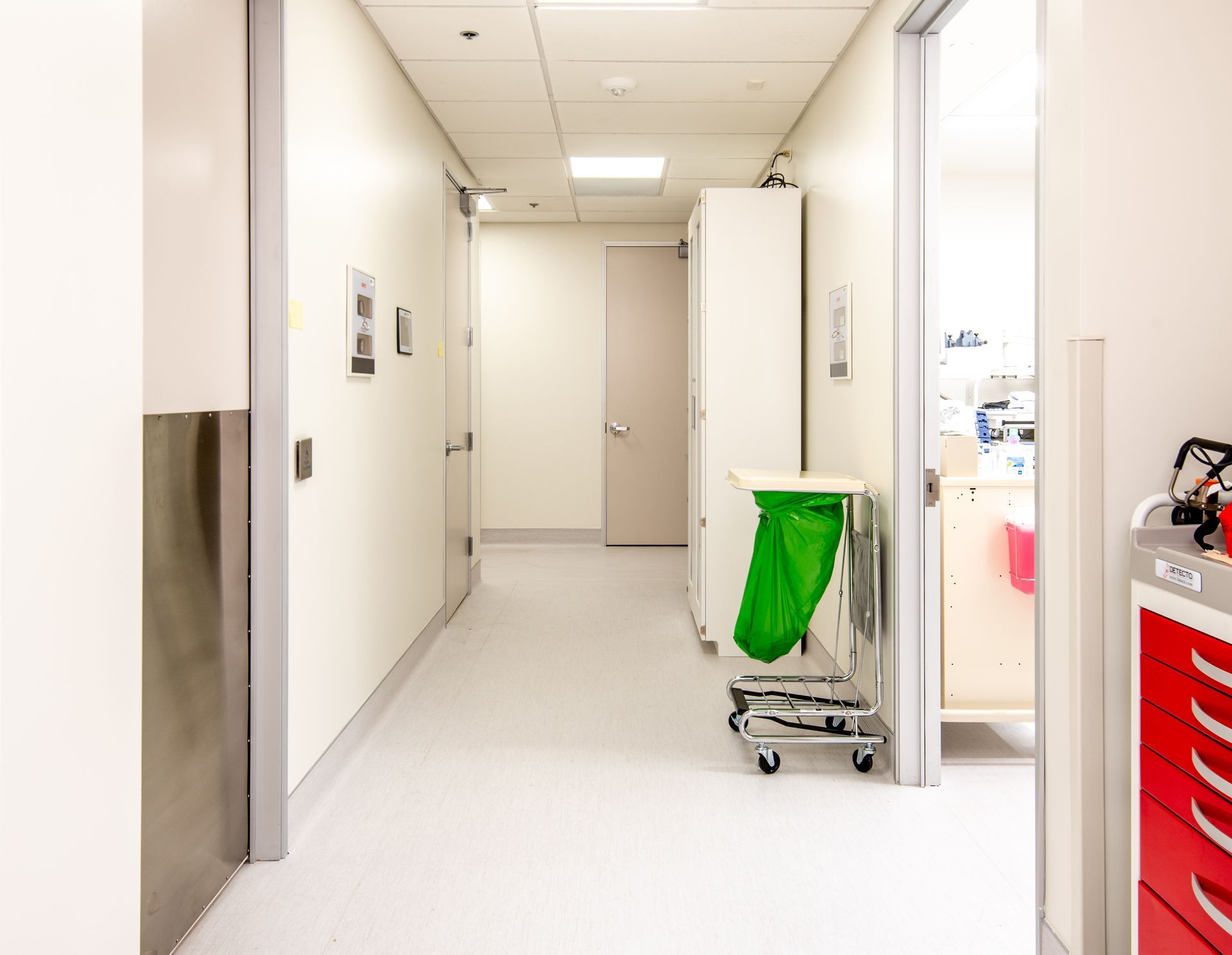 Hallway in a medical facility with white walls, a green waste bin, and a red medical cart.
