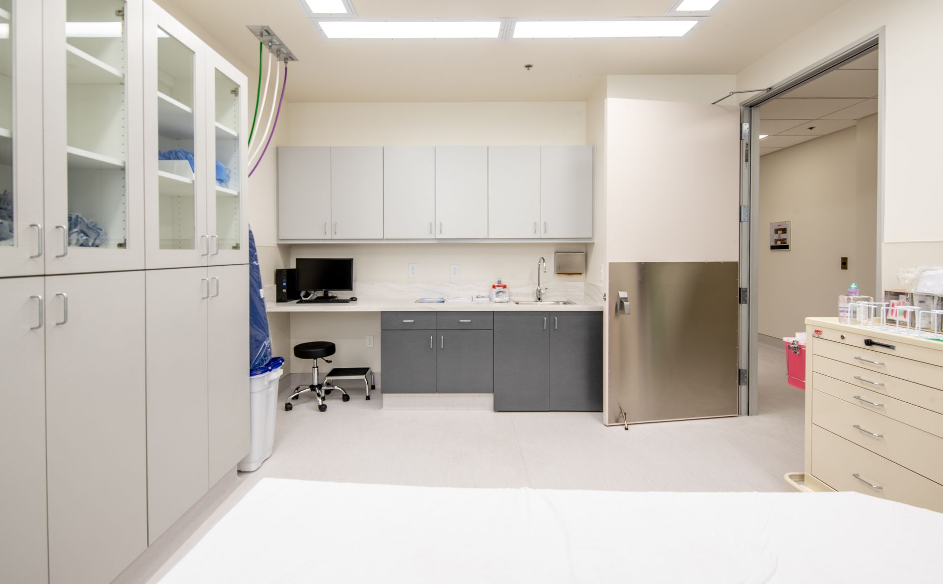 Medical examination room with white cabinets, gray counters, stainless steel sink, and open doorway.