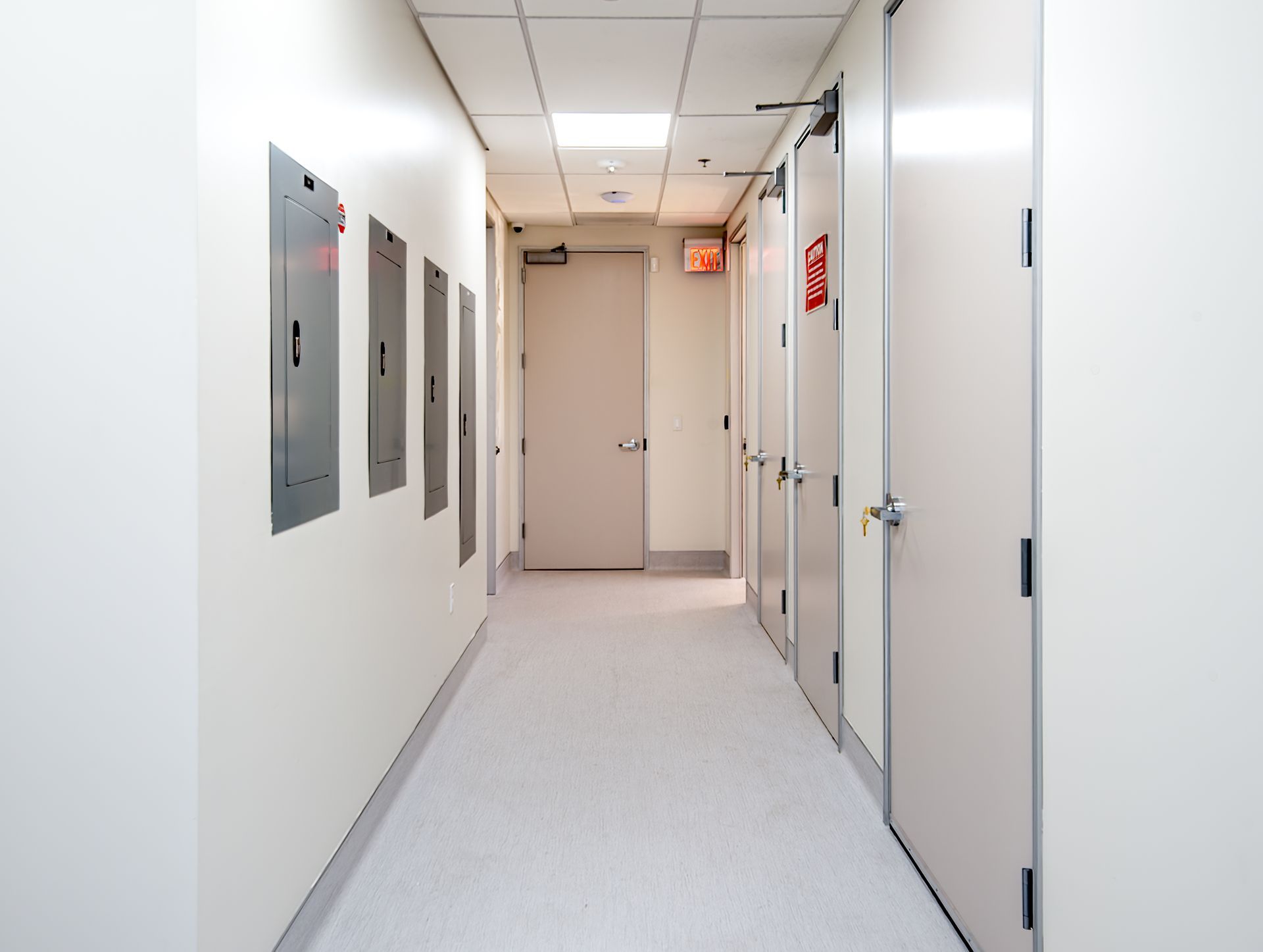 Long hallway with white walls, doors, and ceiling lights. Gray flooring. Electrical panels on the left.