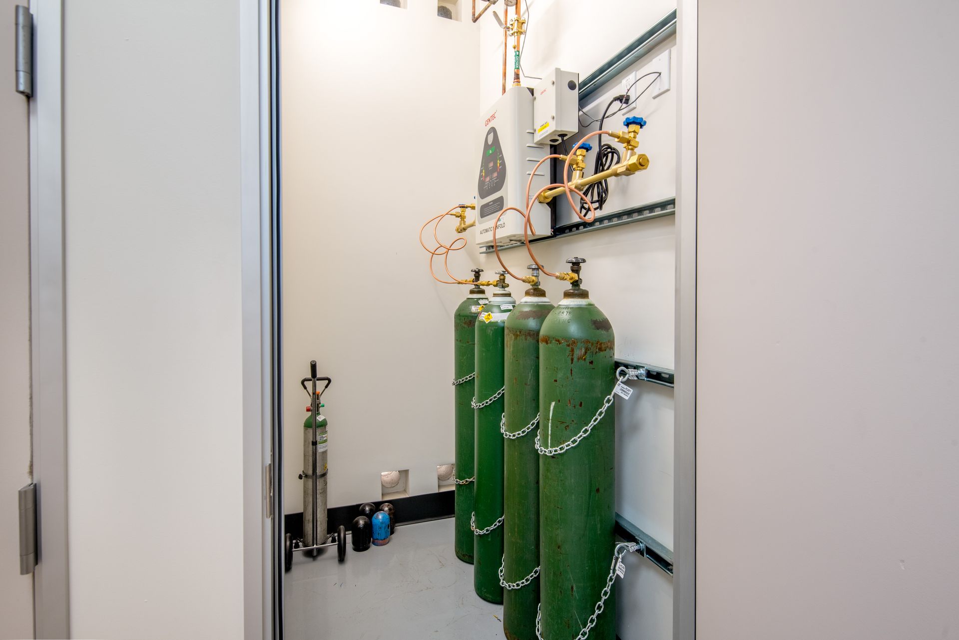 Oxygen tanks chained inside a white utility closet, connected to copper pipes and a panel.