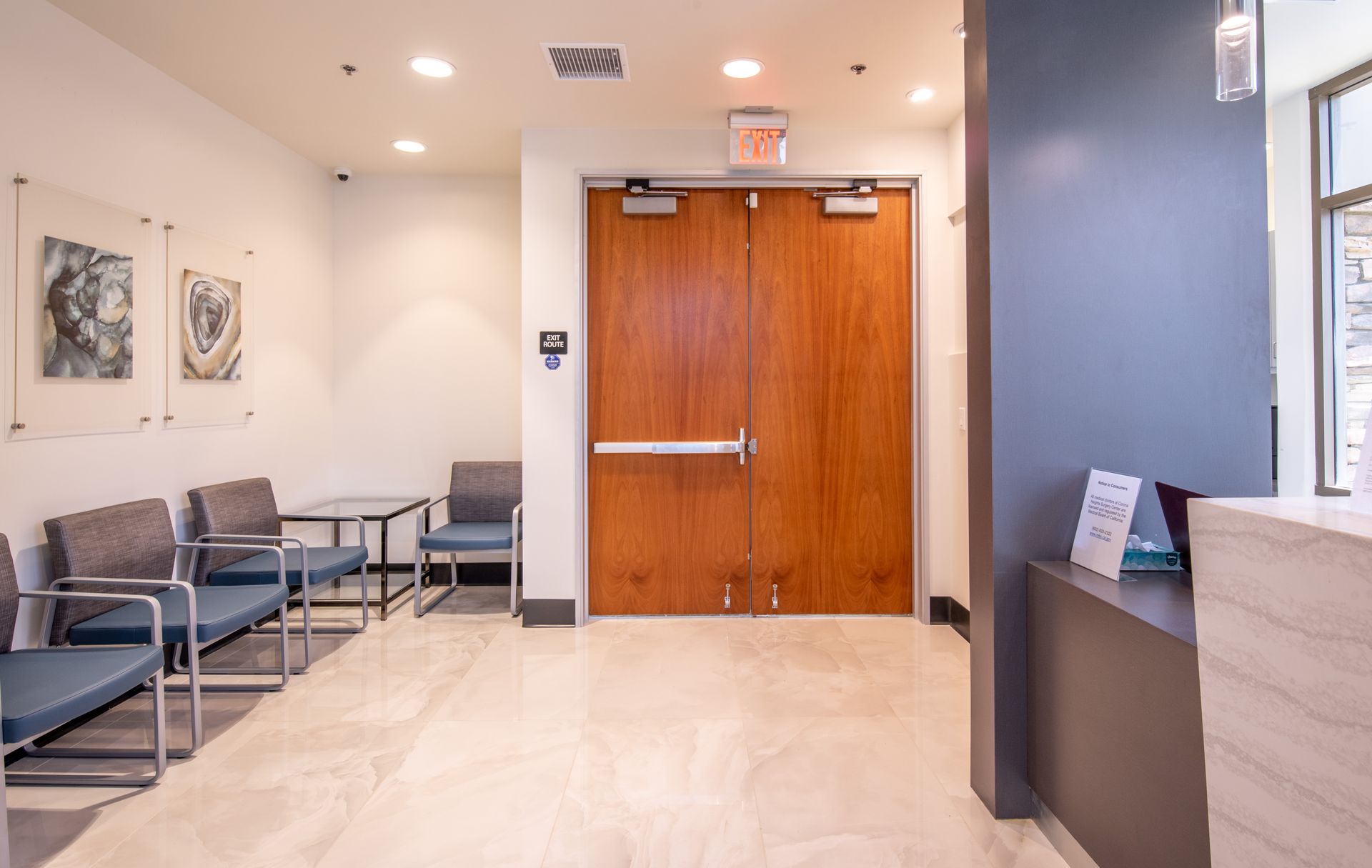 Waiting room with blue chairs, artwork, and wooden double doors leading to an exit.
