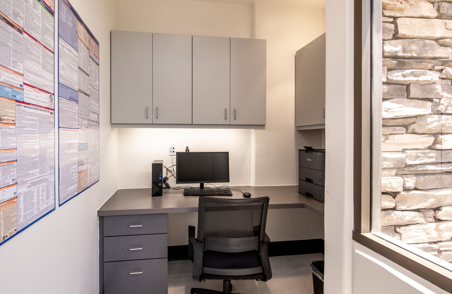 Office nook with desk, computer, chair, cabinets, and stone wall visible through window.