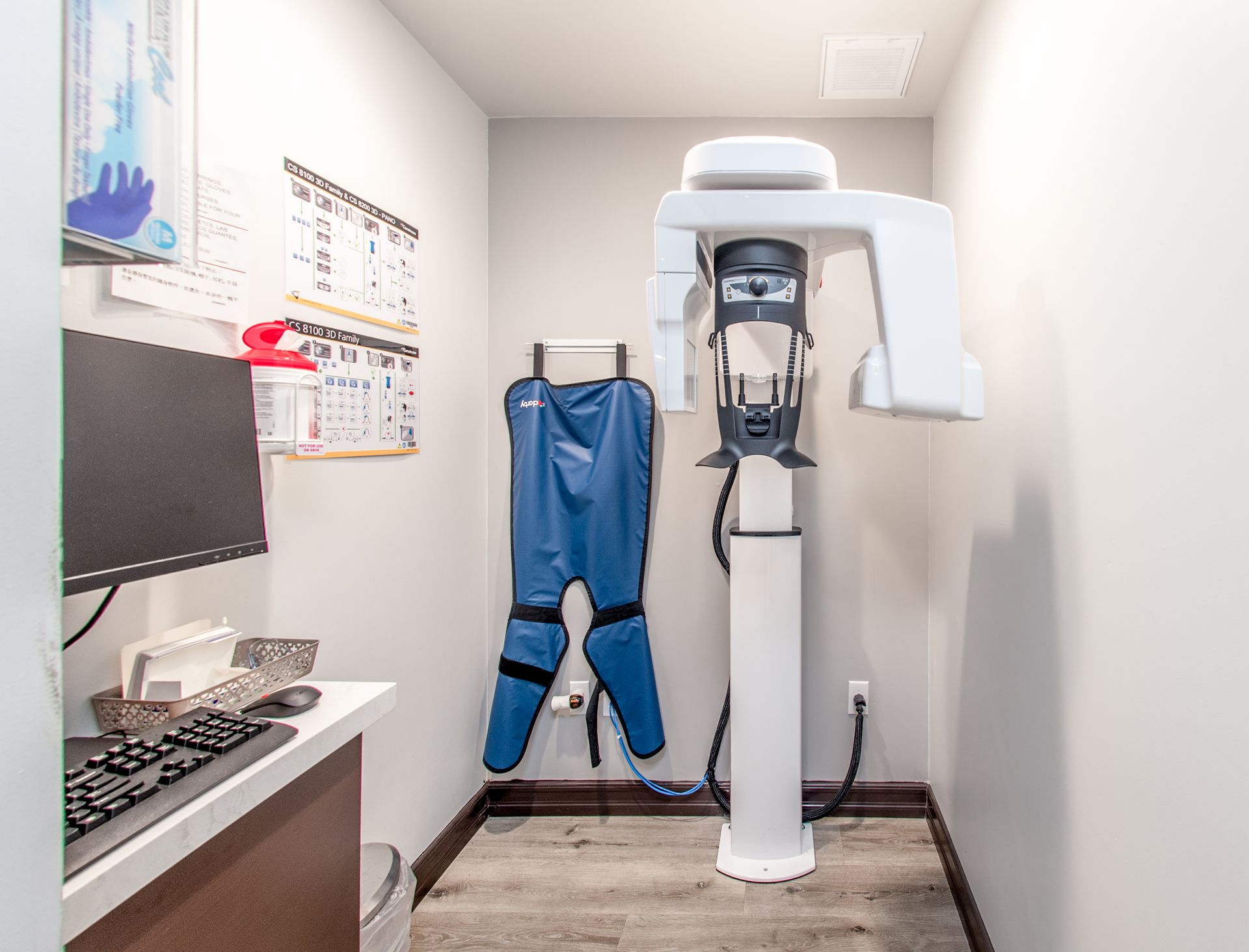 A dental X-ray room with a machine, lead apron, computer, and charts.