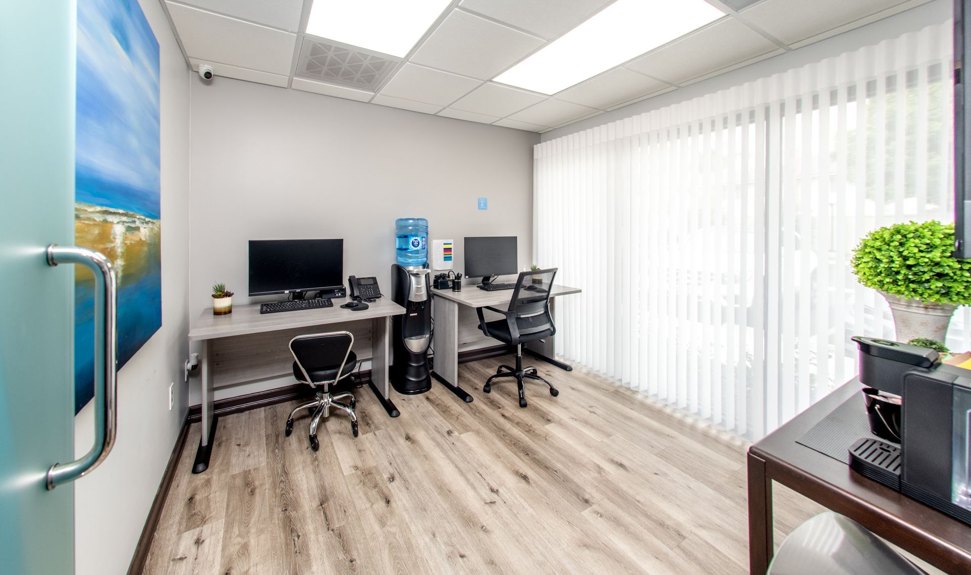 Office space with two desks, computers, and water cooler, window with blinds, and a small plant.