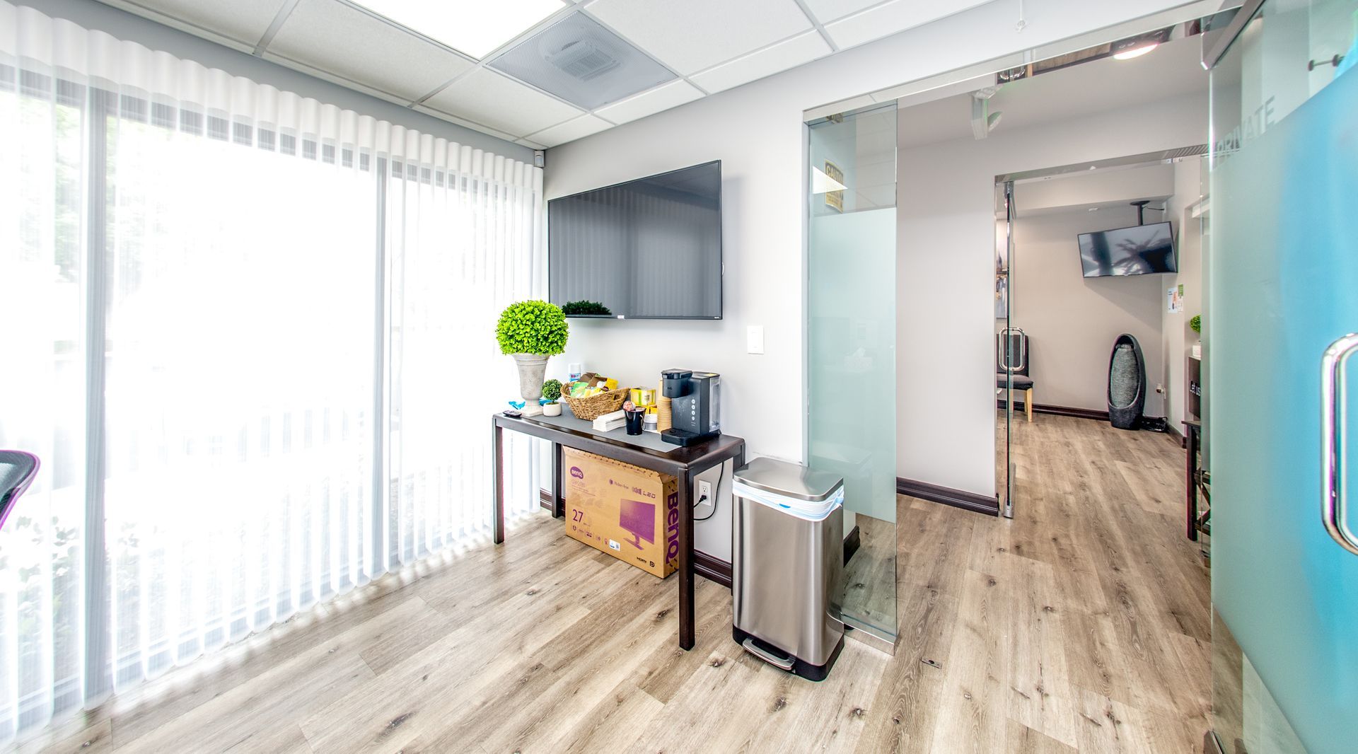 Office waiting area with TV, refreshments, and a window. Light wood floor, glass door.