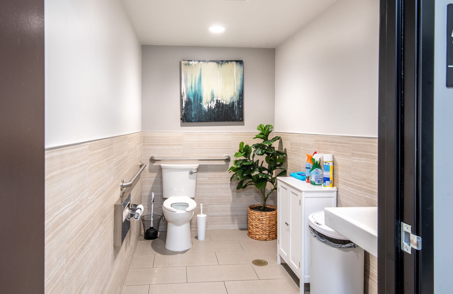 Accessible public restroom with toilet, sink, grab bars, and art; neutral tones and a plant.