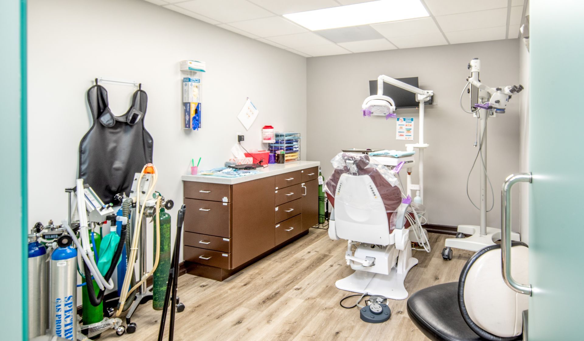 Dental exam room with chair, equipment, and supplies. Light wood floor, neutral walls, and a partial view of the door.