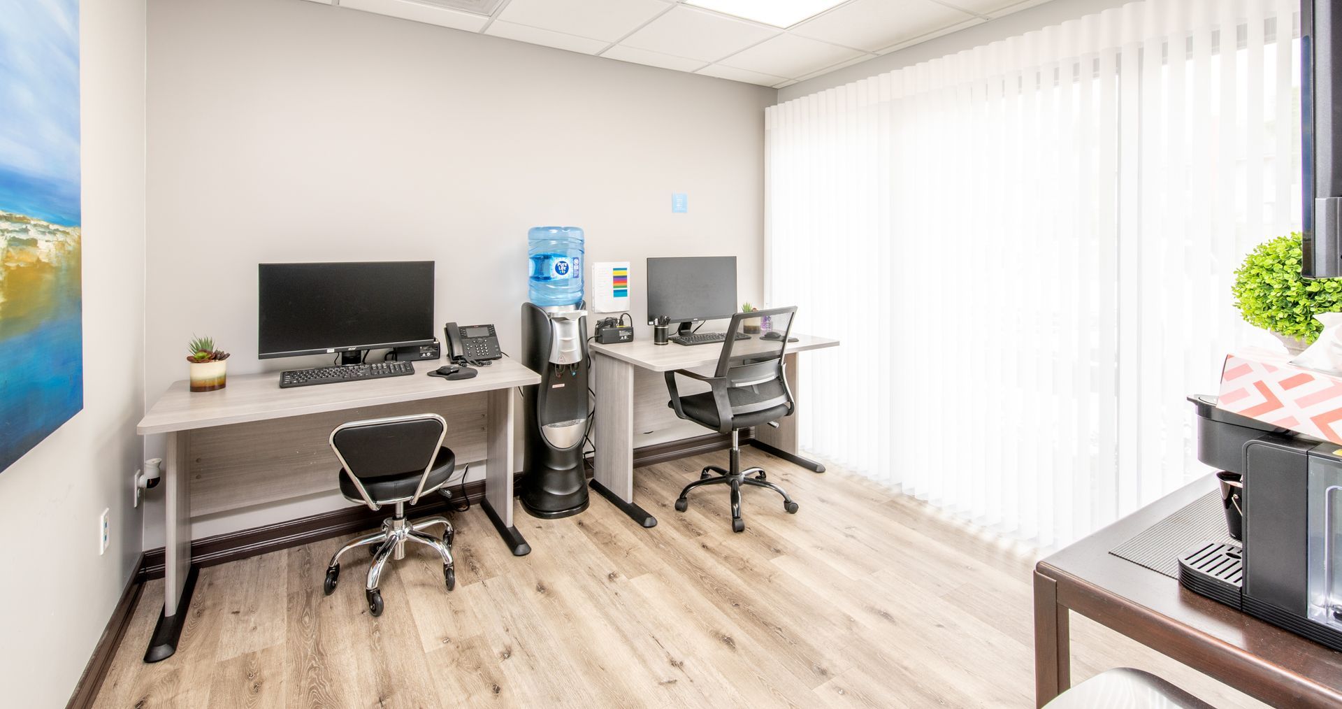 Two office desks with computers, water cooler, and white vertical blinds.