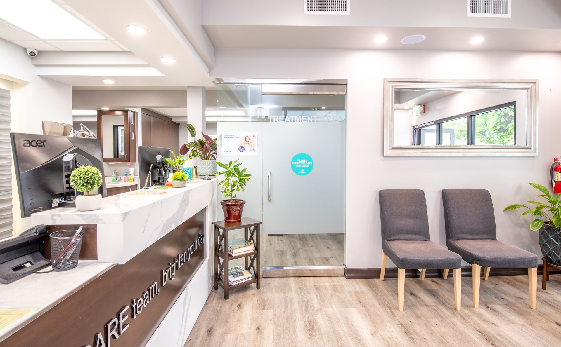 Reception area with desk, waiting chairs, plants, and frosted glass door with logo.