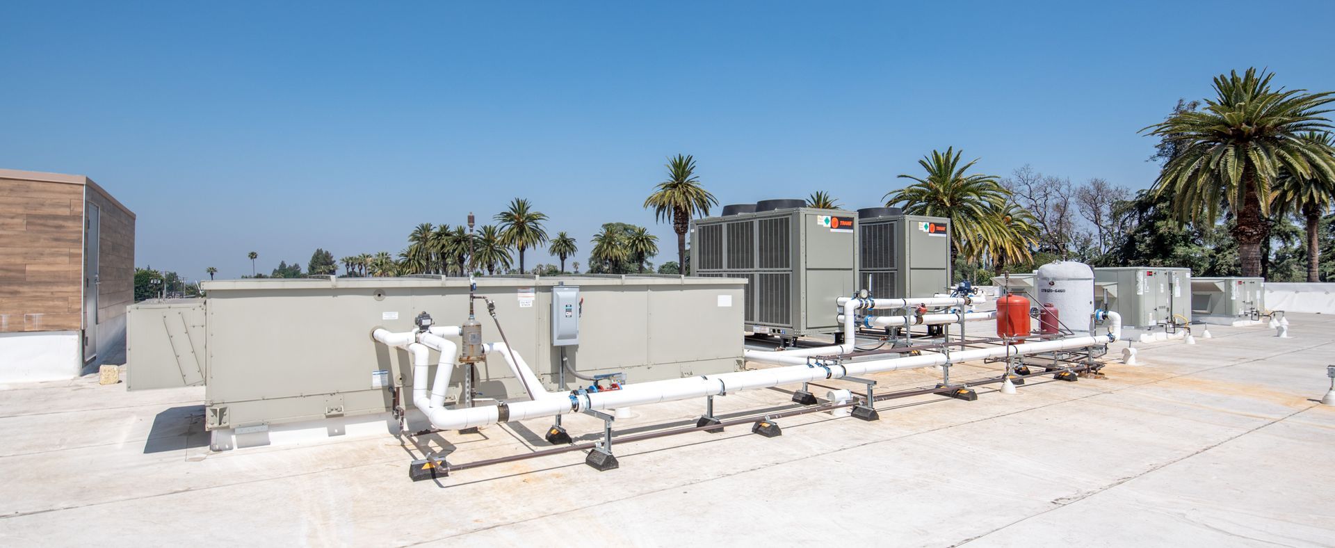 Rooftop HVAC system with pipes, tanks, and palm trees against a clear blue sky.